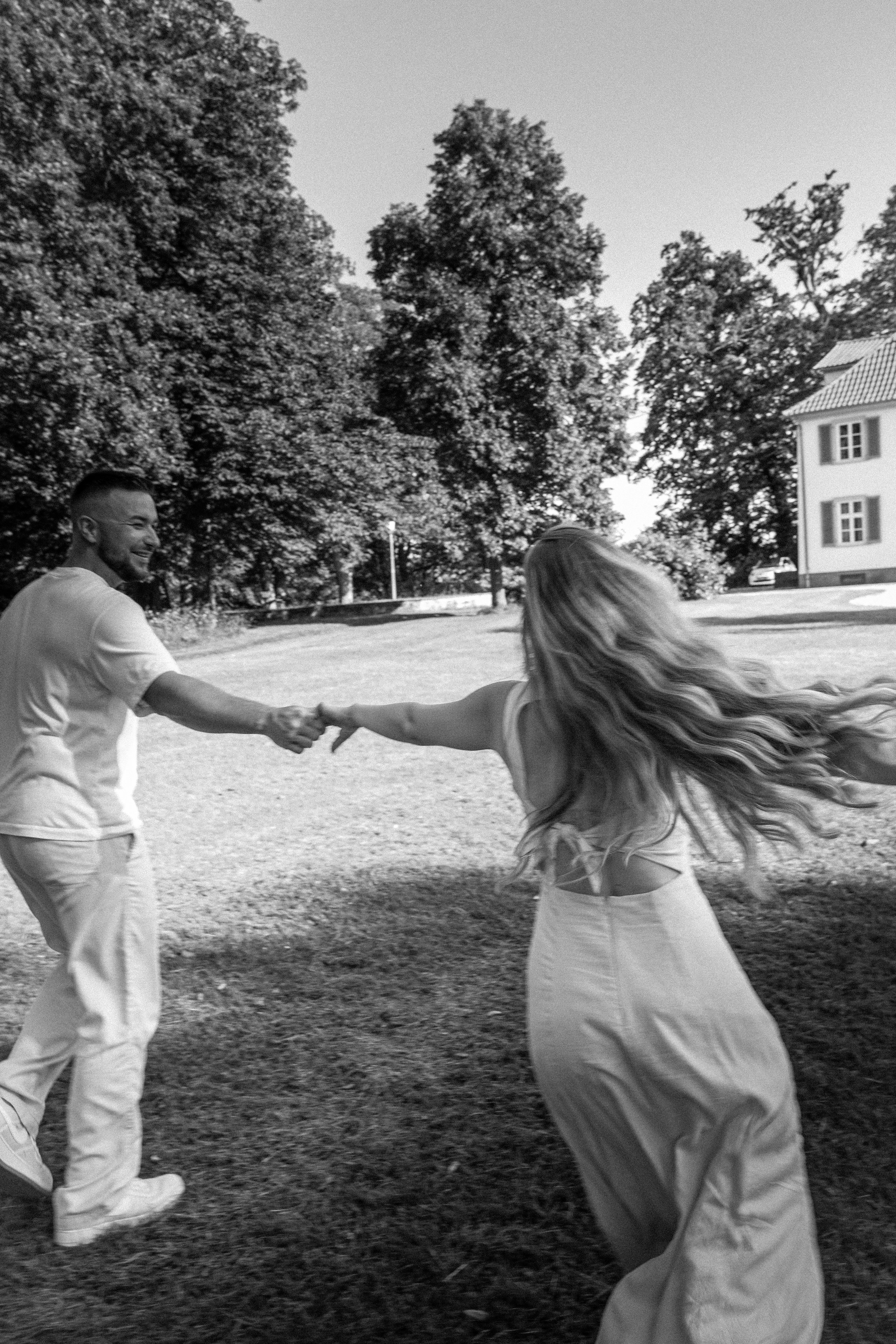 Zwei Menschen, die im Park tanzen, während es sonnig ist. Einer ist ein Mann, der in einem weißen T-Shirt und weißen Hosen gekleidet ist, die andere Person ist ein Mädchen mit langen, gewellten Haaren und einem weißen Kleid.