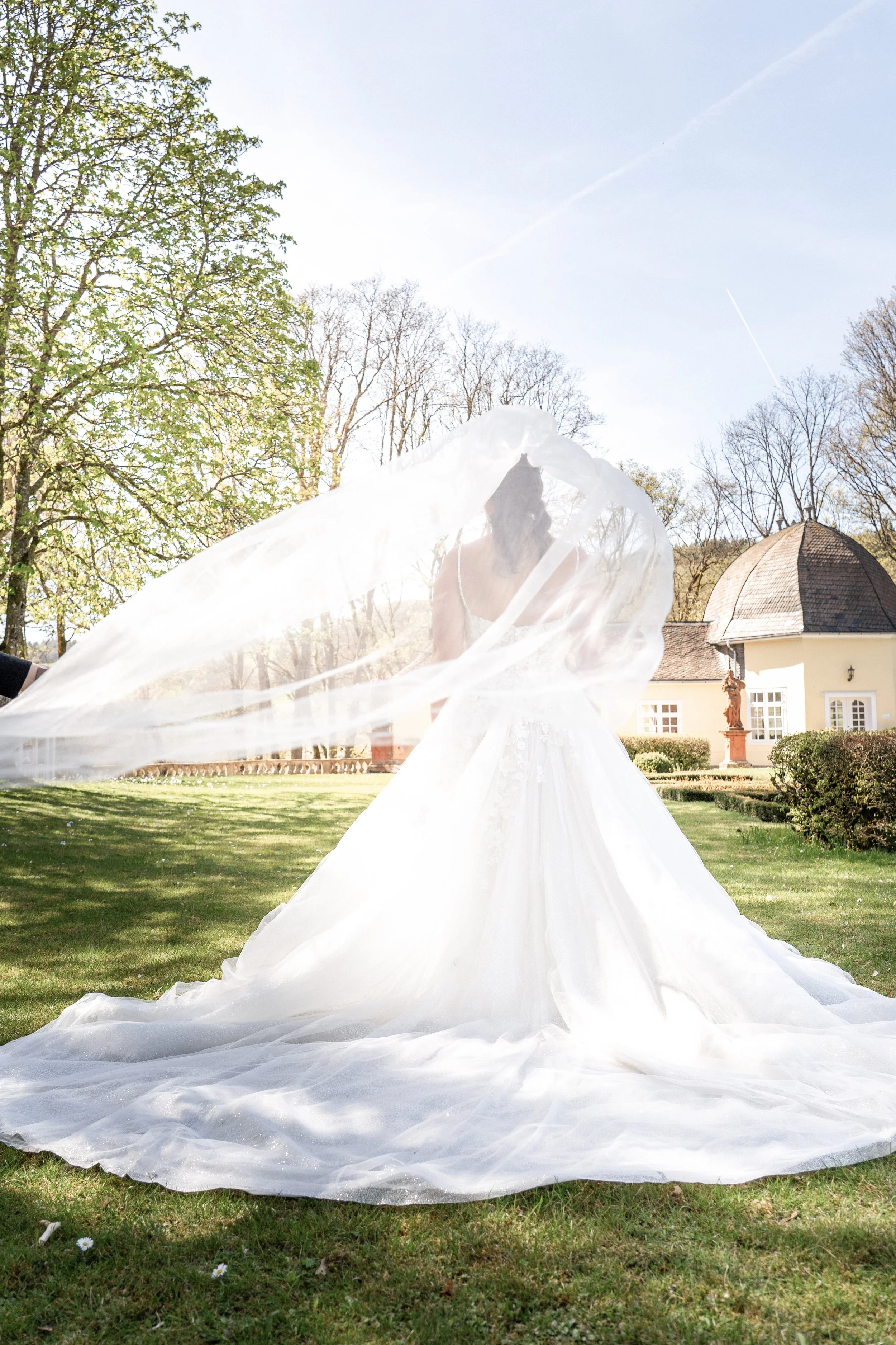 Eine Frau in einem weißen Brautkleid, die im Freien auf einer Wiese steht. Sie hat einen langen, schimmernden Schleier, der sich auf dem Boden ausbreitet. Im Hintergrund sind Bäume, ein Gebäude mit Kuppeldach und eine Statue zu sehen, bei sonnigem Hi