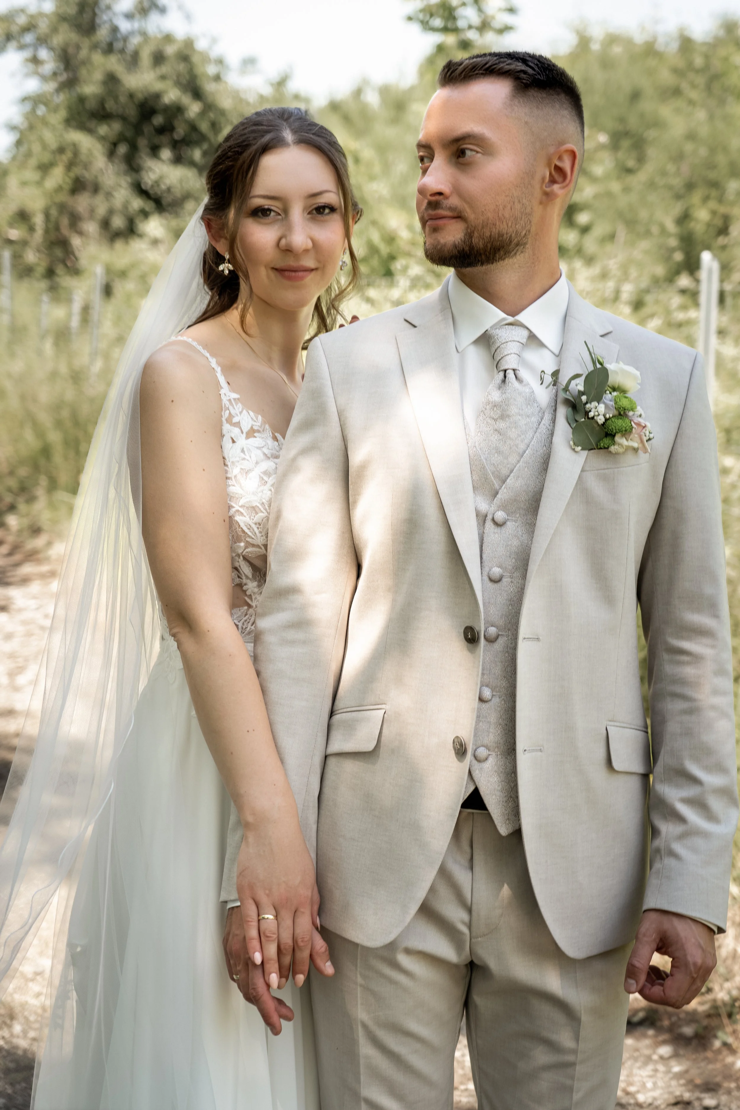 Ein Brautpaar bei einer Hochzeit im Freien, die Braut trägt ein weißes Kleid mit Spitze, der Bräutigam trägt einen hellen Anzug mit Krawatte. Sie halten sich an den Händen. Im Hintergrund sind Bäume und ein blauer Himmel sichtbar.