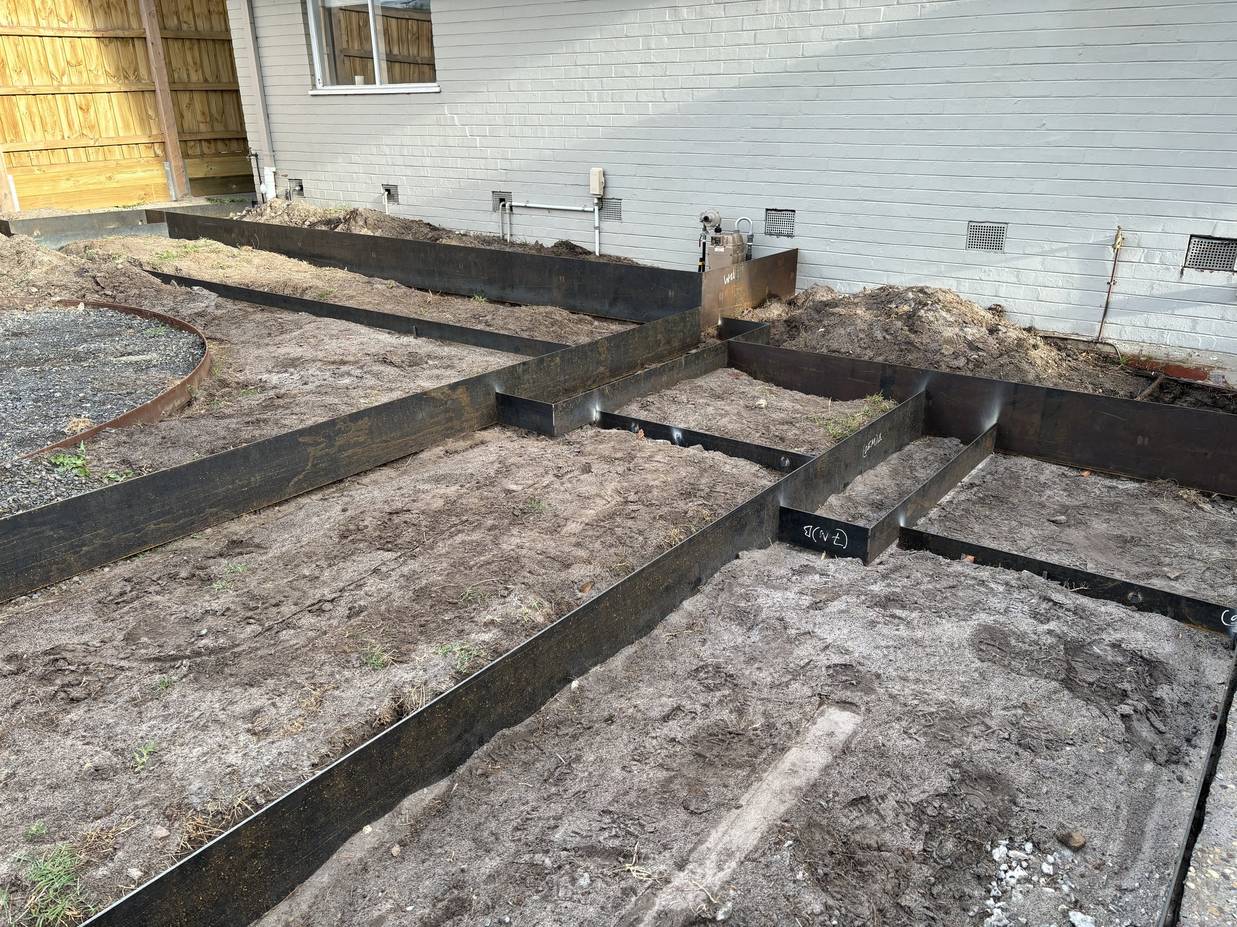 Construction site with black metal framing for a house foundation, dirt ground, and a brick house wall in the background.