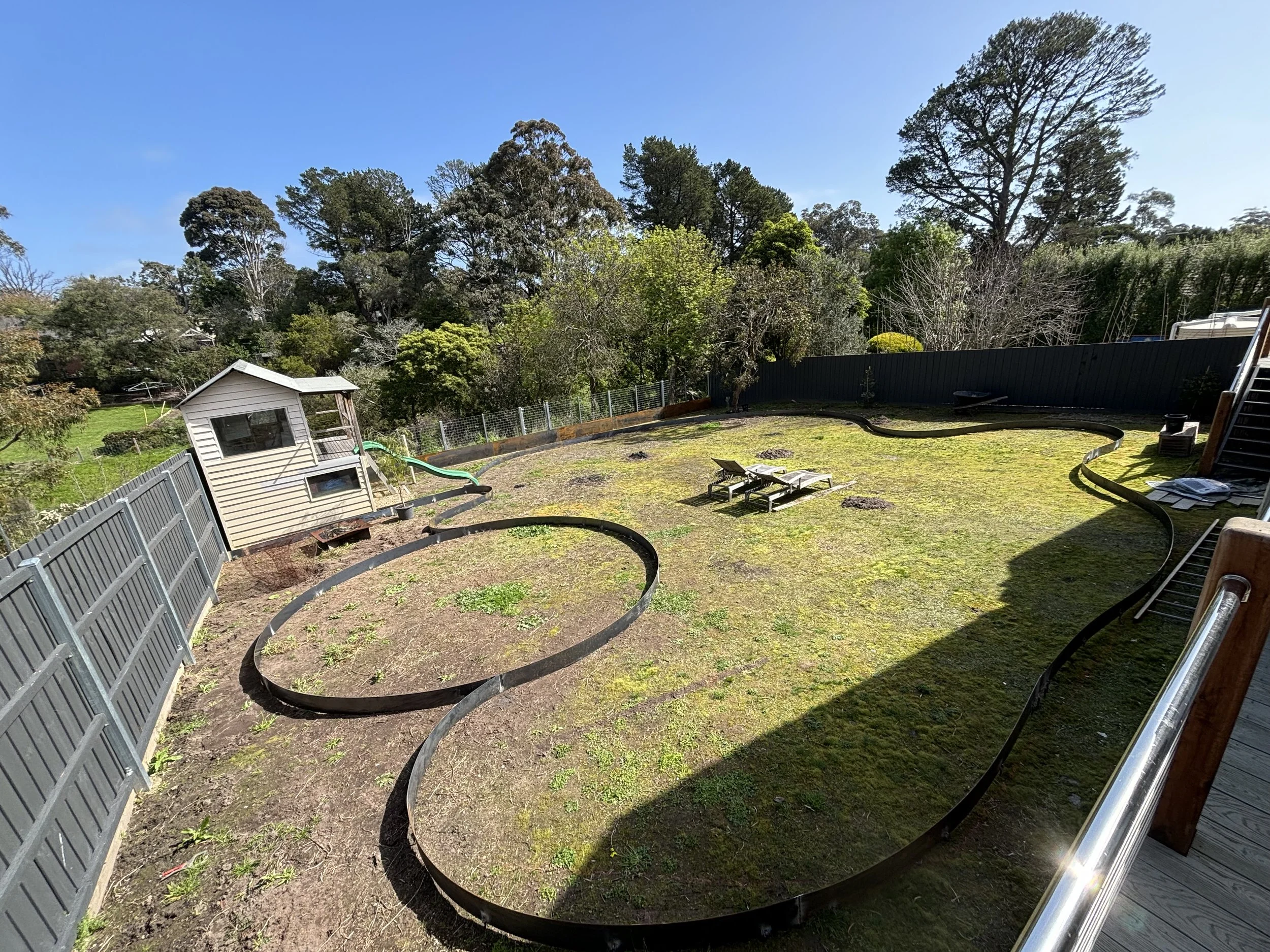 A backyard with a small white shed, a fence, trees, and a slide, showing a partially tilled patch of soil and some outdoor furniture under a blue sky.