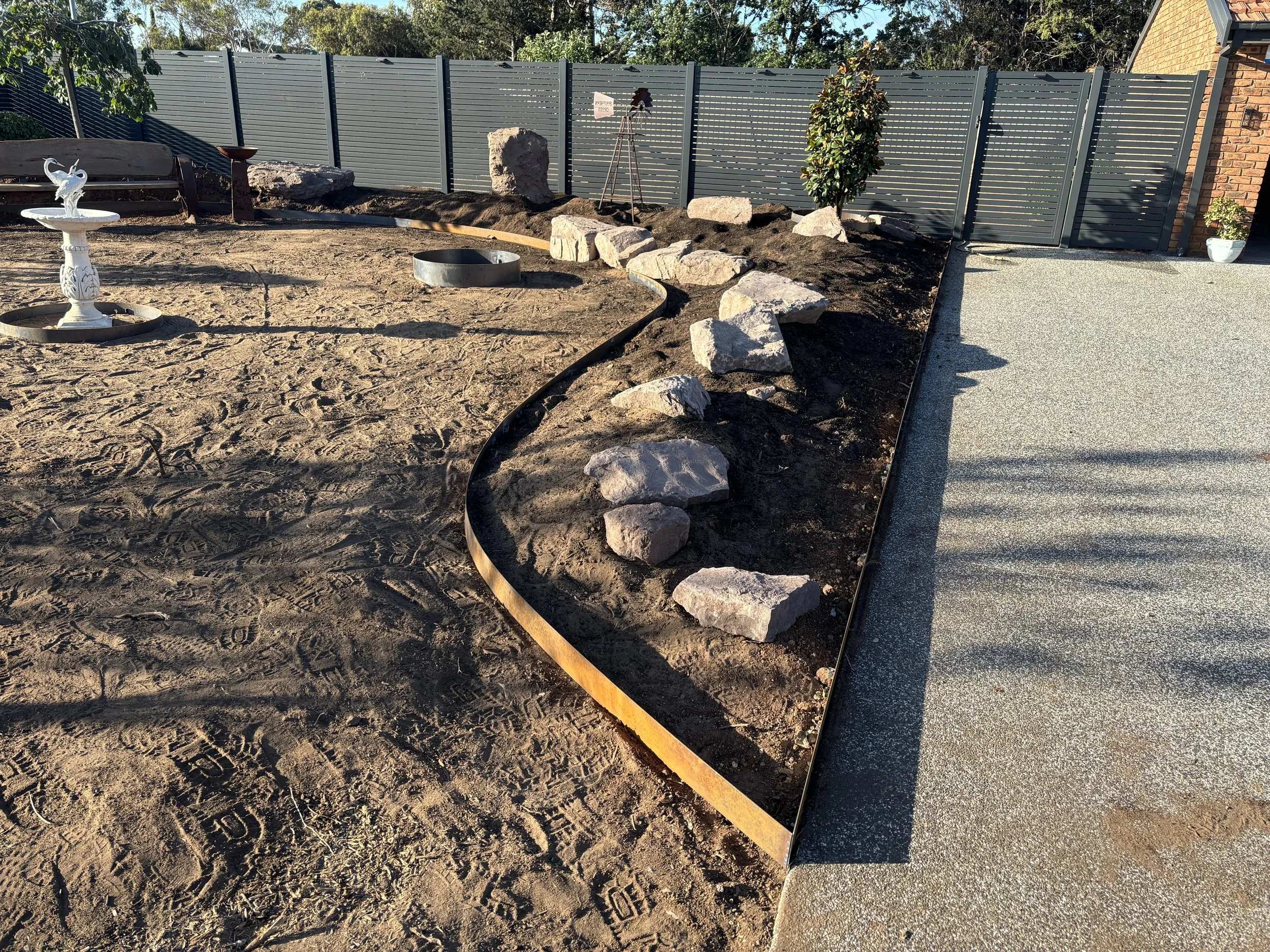 A backyard landscape under construction with a curved steel and corten edging  line separating large rocks and a dirt area from a paved surface