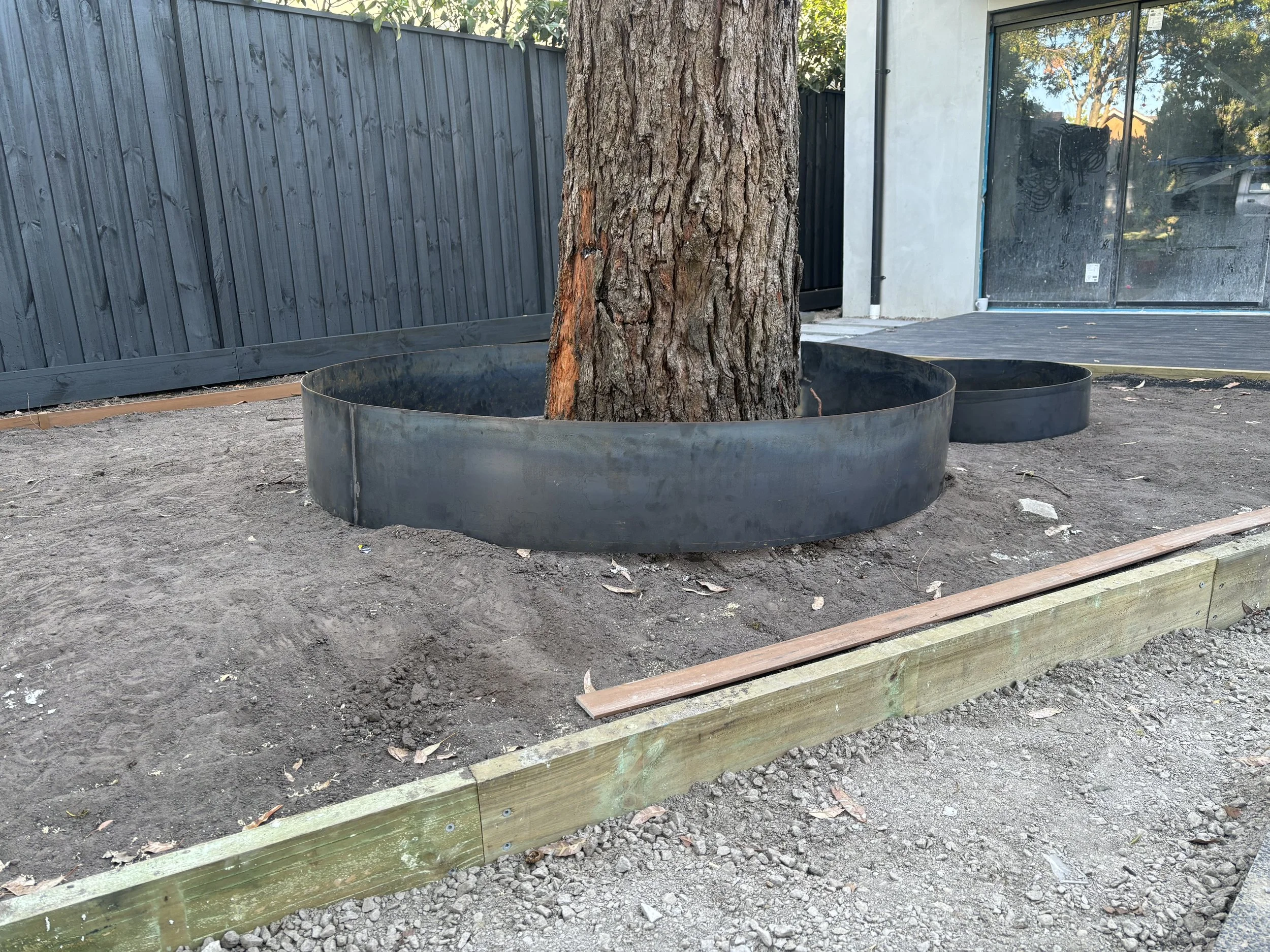A tree planted in the ground within a large, black, circular steel and corten barrier