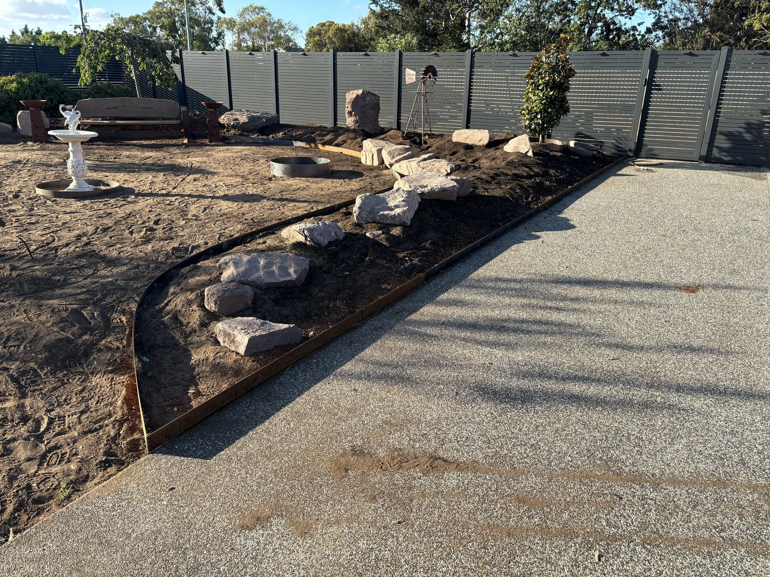 A landscaped backyard area with a gravel pathway, a planted area with large rocks, a decorative bird bath