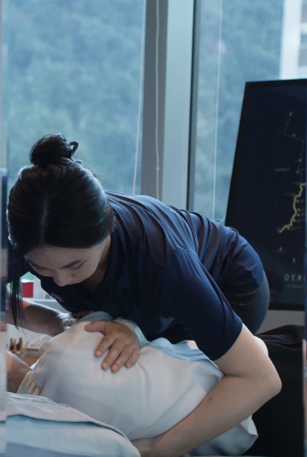 A woman performing CPR on a person lying on a hospital bed with a monitor displaying medical data in the background.