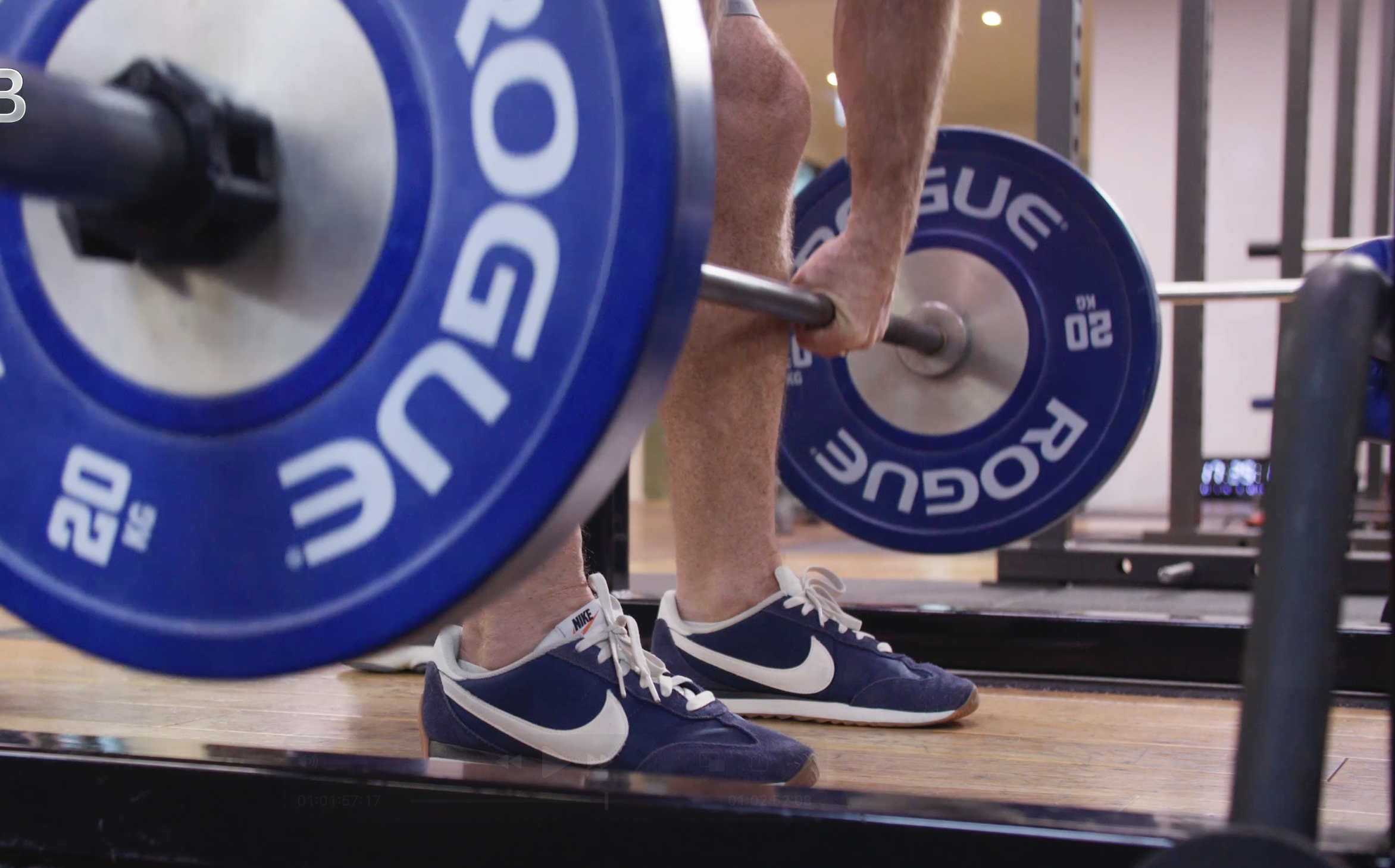 Close-up of a person lifting a blue barbell plate labeled '20 KG' during weightlifting, with another plate visible in the background, and the person's legs and sneakers in focus.