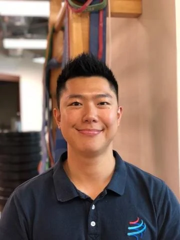 Portrait of a smiling young man with short black hair, wearing a dark blue polo shirt with a logo on the chest, standing indoors near a wooden wall with exercise resistance bands hanging in the background.