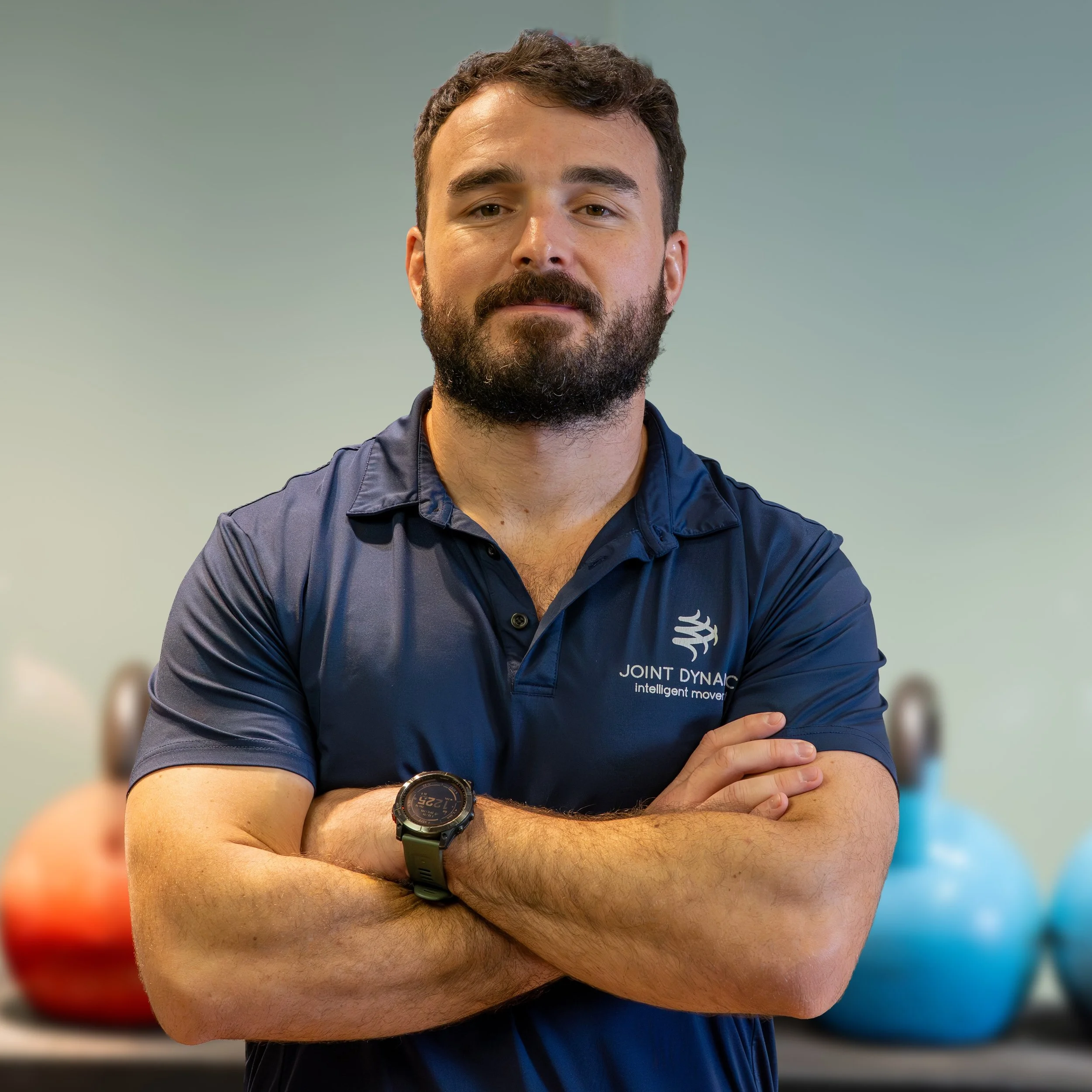 A man standing in a gym or fitness center, smiling, wearing a navy polo shirt with a logo, with workout equipment and wooden flooring in the background.