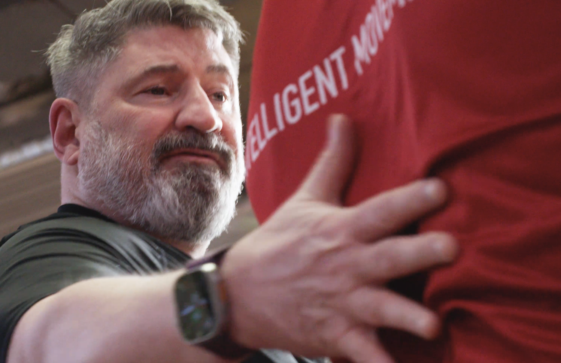 A man with gray hair and a beard touches another person's red shirt, which has white text on it, with a concerned or serious expression on his face.
