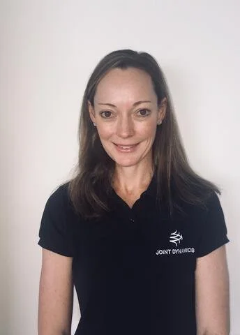 A woman with long brown hair wearing a black shirt with a logo and the words 'Joint Dynamics' printed on it, standing against a white background.