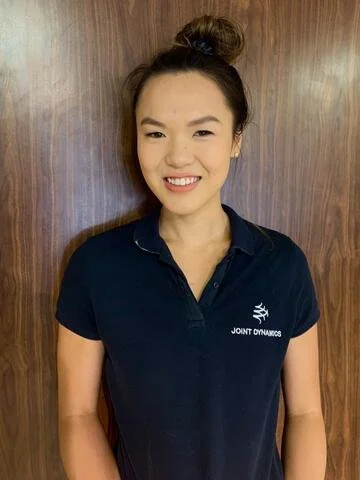 A young woman with dark hair tied up in a bun, smiling, wearing a navy blue polo shirt with a logo that reads "Joint Dynamics," standing against a wooden wall.