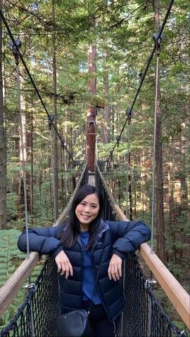 A woman standing on a suspension bridge in a forest, smiling, surrounded by tall trees.