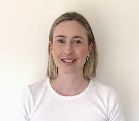 A young woman with blonde hair, wearing a white shirt and pearl earrings, smiling at the camera in front of a plain white wall.