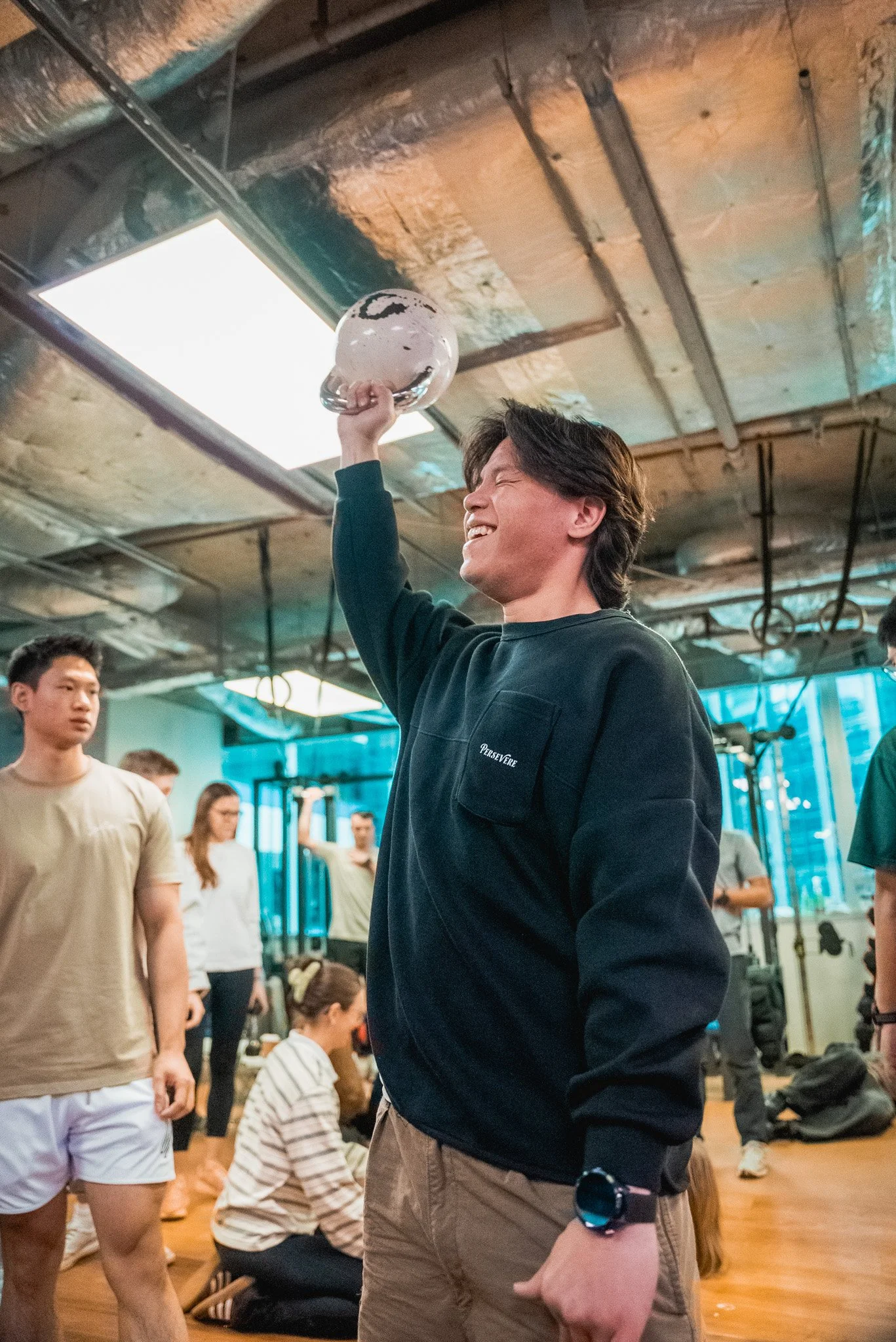 A young man with dark hair smiling and holding a kettlebell in a gym or fitness studio, surrounded by other people, some of whom are kneeling or standing, with exercise equipment and mirrors in the background.
