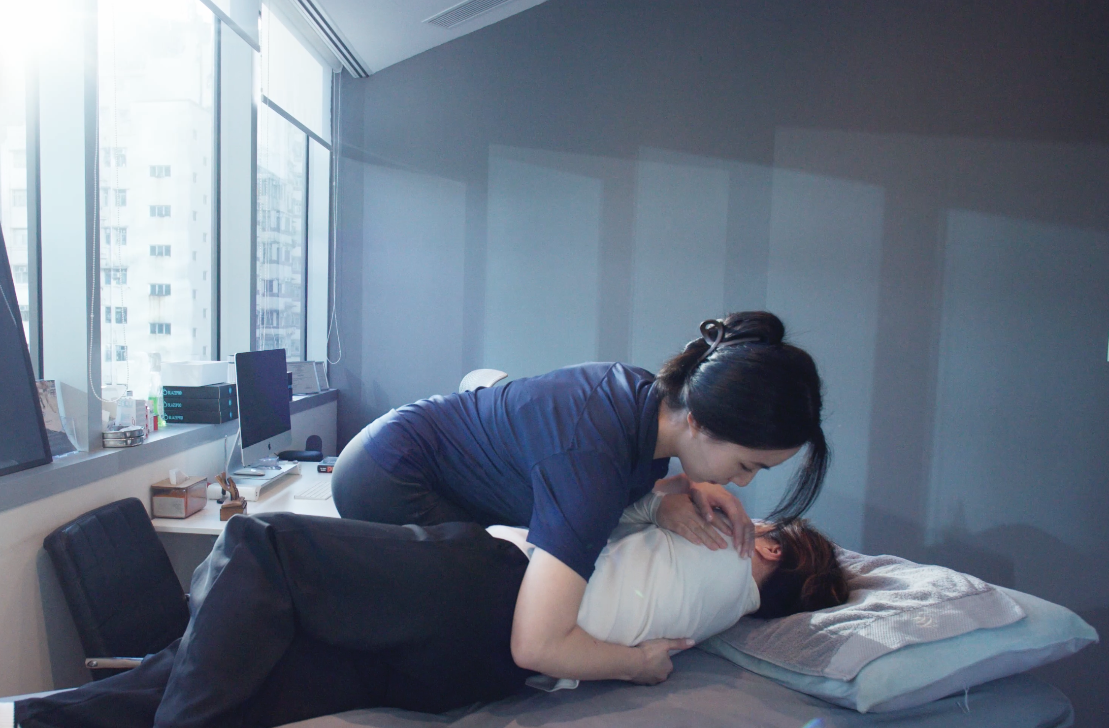 Two people performing CPR on a person lying on a bed in an office space with large windows.
