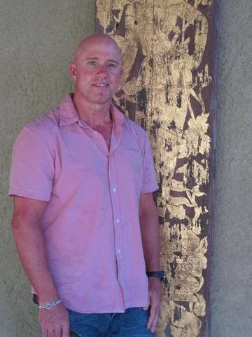 A man with a shaved head wearing a pink short-sleeve button-up shirt and blue jeans standing next to a textured wall with a large gold and black artwork.