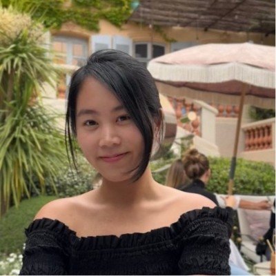 Young woman with short black hair wearing an off-the-shoulder black top, standing outdoors near a garden with umbrellas and someone in the background.