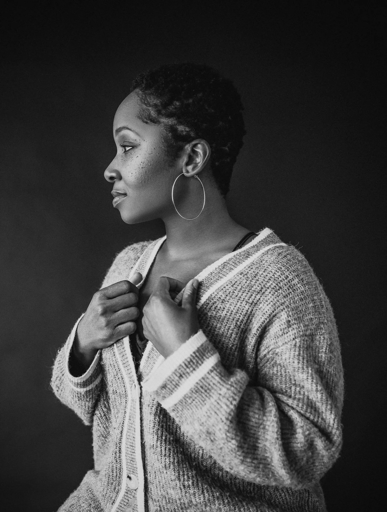 A black and white portrait of a woman with short, textured hair, wearing large hoop earrings and a cozy, striped cardigan, looking to the left.