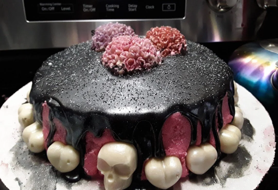 A gothic-themed cake decorated with black icing, white skull-shaped candies around the base, and pink frosting flowers on top, with a sprinkle of silver edible glitter on the surface.