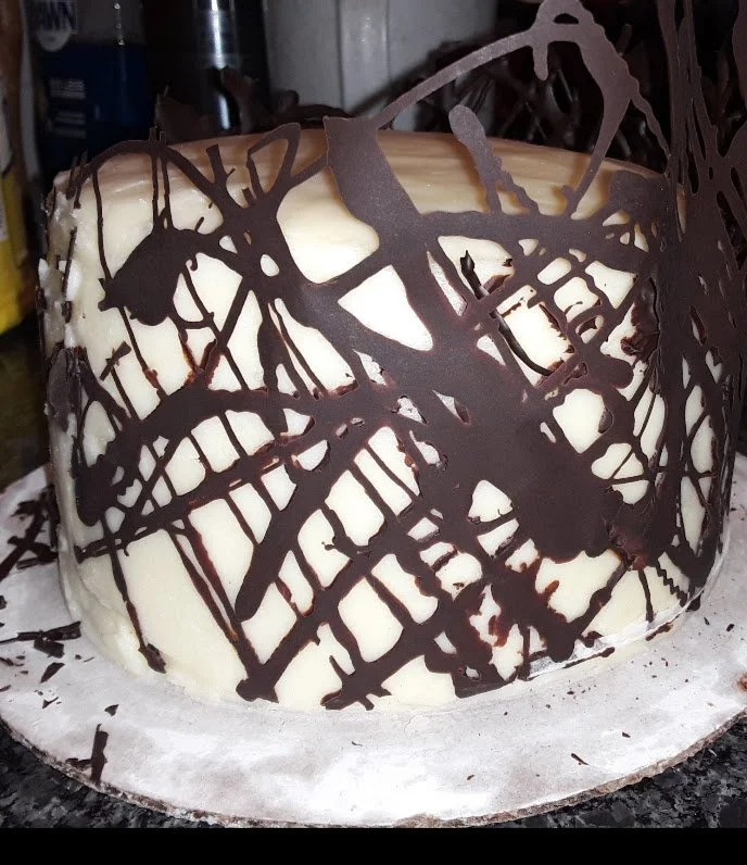 A round cake with white frosting decorated with dark chocolate drizzle in an abstract pattern.
