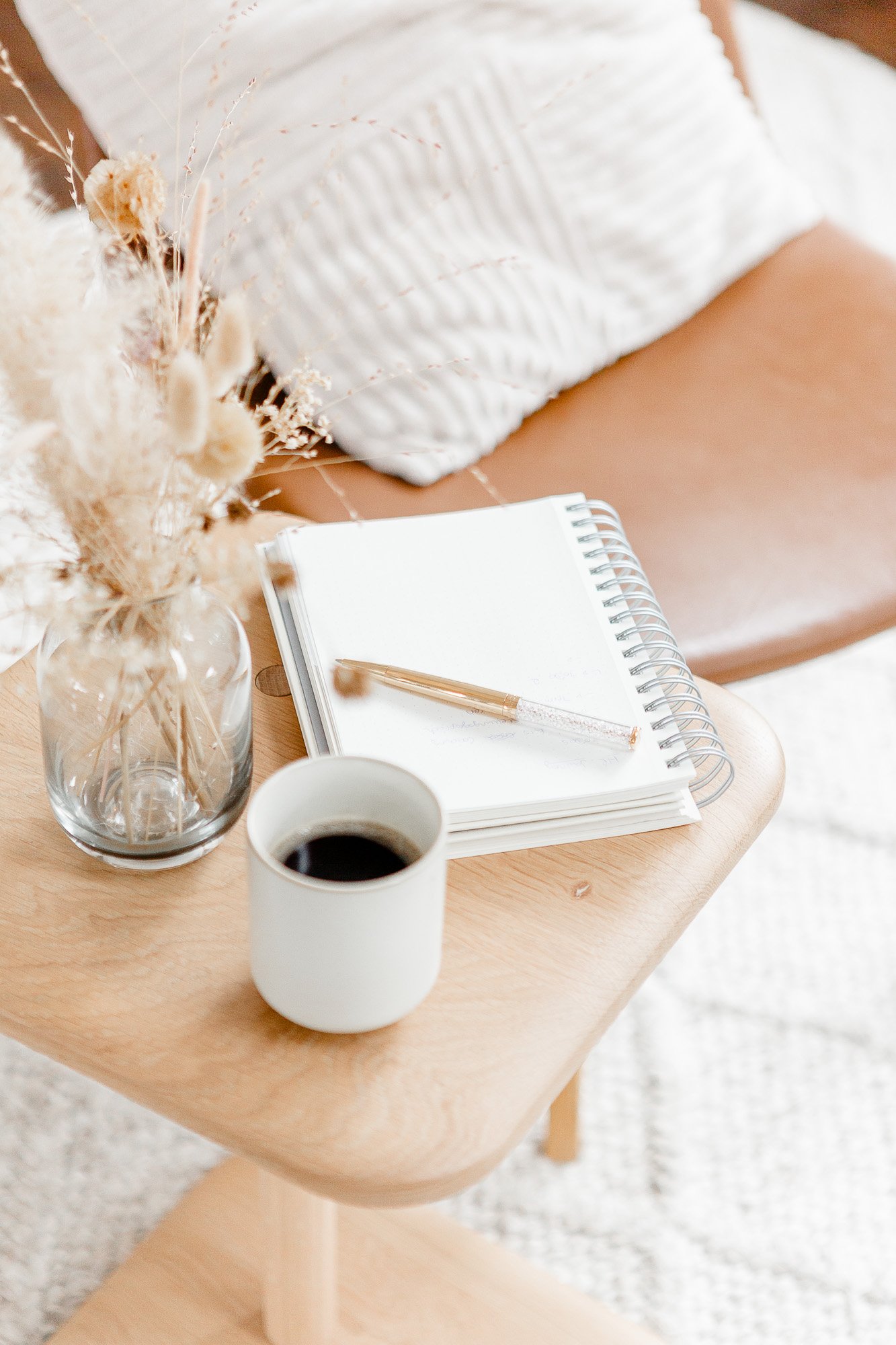 Eine Tasse Kaffee, ein Notizbuch mit einem Kugelschreiber, eine Vase mit getrockneten Blumen auf einem kleinen Holztisch, im Hintergrund ein Sofa mit weißen Kissen.