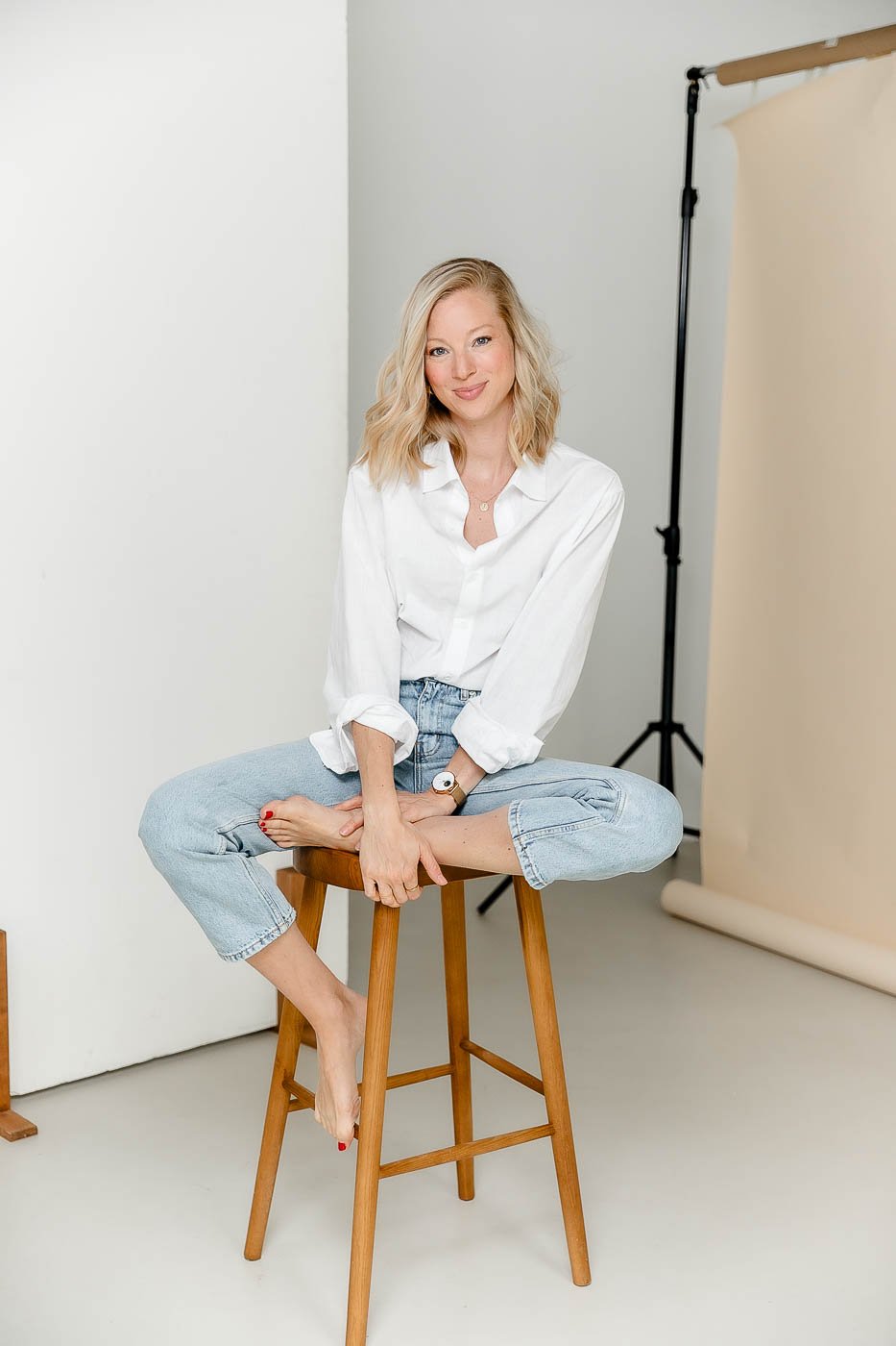 Eine junge Frau sitzt auf einem Holzhocker, die Beine kreuzt. Sie trägt eine weiße Bluse und Jeans, hat blonde, lockige Haare und lächelt. Im Hintergrund steht ein Tonstudio-Ständer mit einem beigefarbenen Hintergrund.