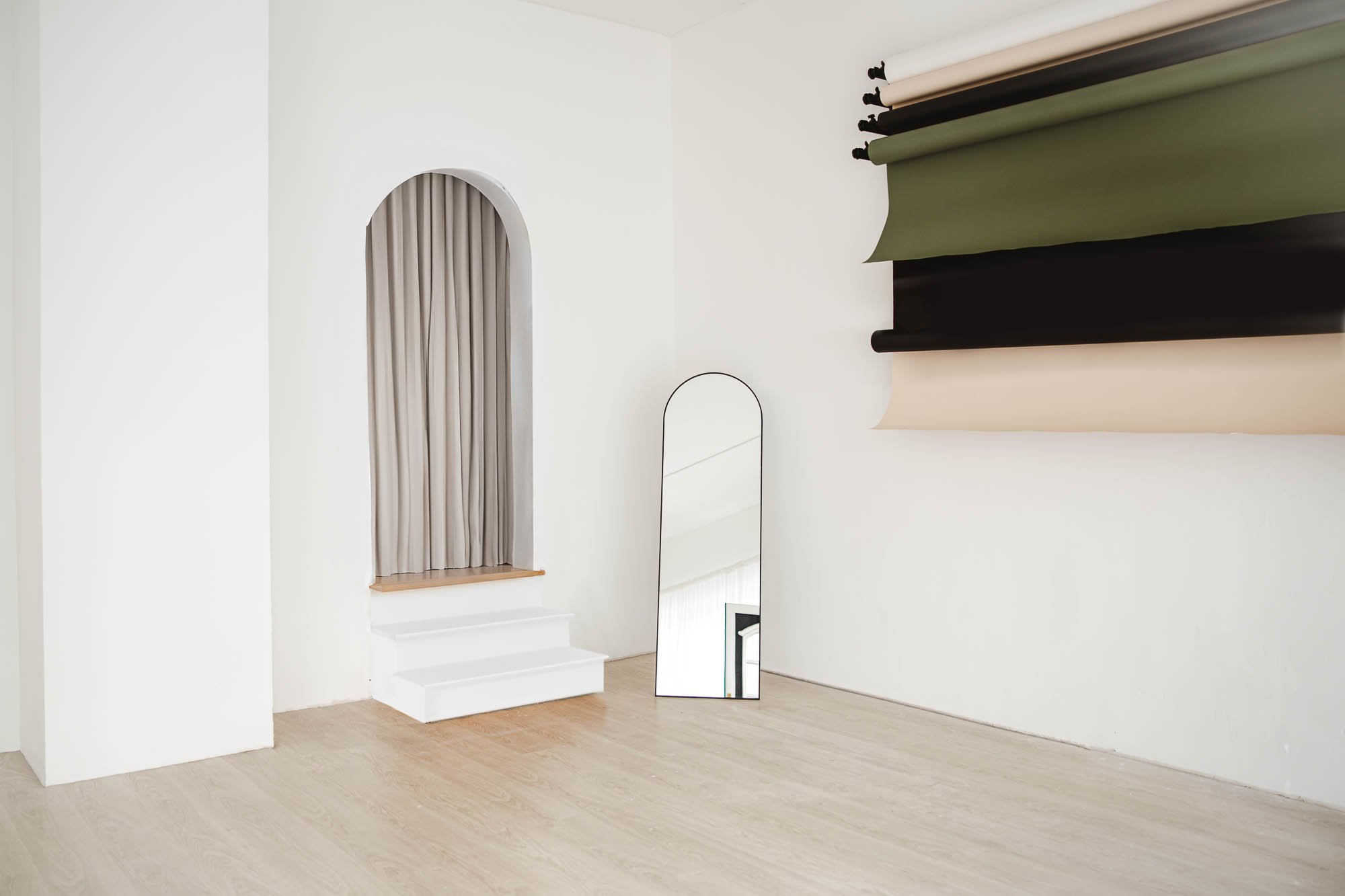 Ein leerer Raum mit weißen Wänden, Holzboden, einem großen Spiegel und mehreren aufgerollten Stoffbahnen an der Wand