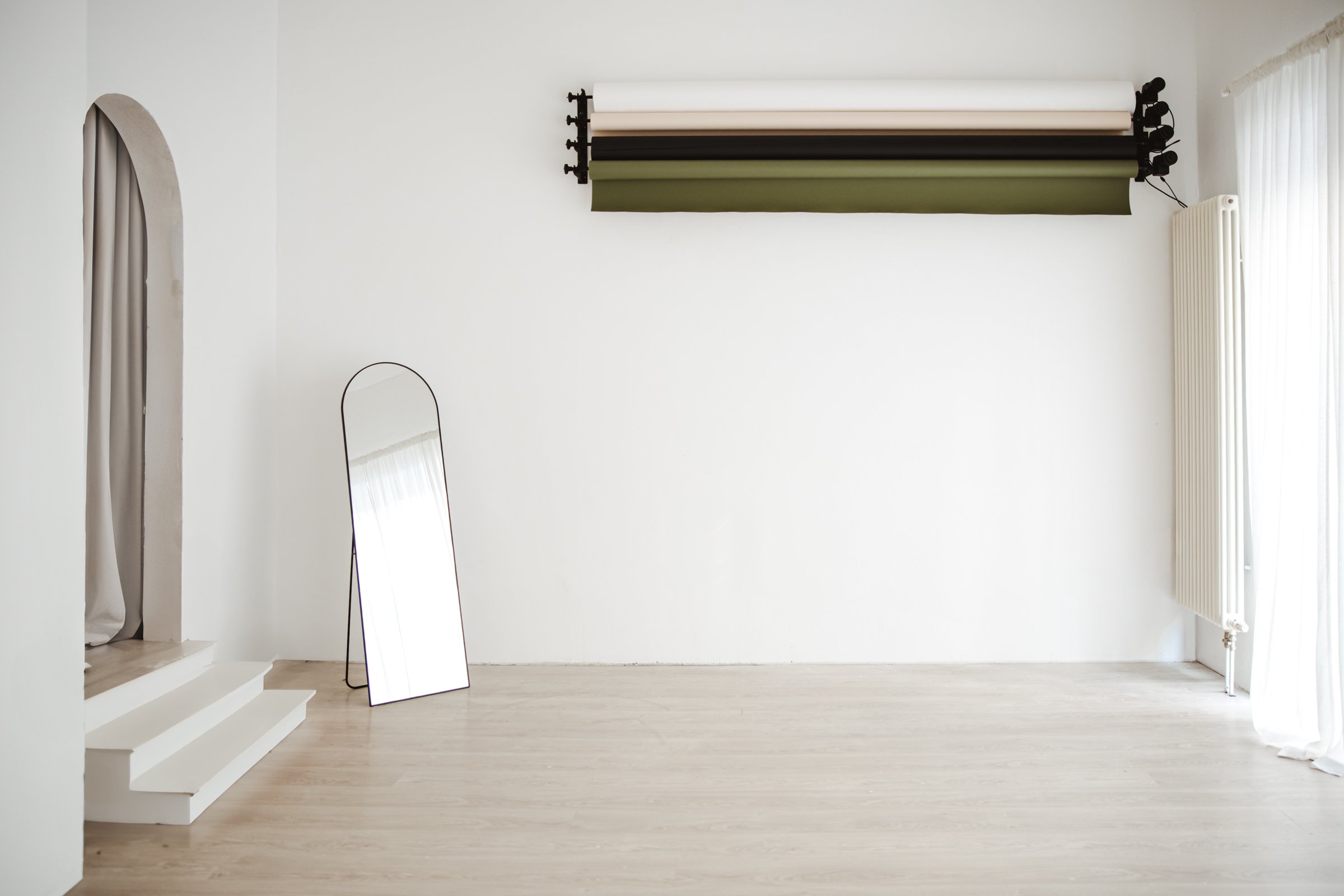 Mietstudio mit automatischem Hintergrund-Rollen-System an der Wand mit 4 Farbrollen in. weiß, beige, olivgrün und schwarz sowie großem Standspiegel und ästhetischem Rundbogen.