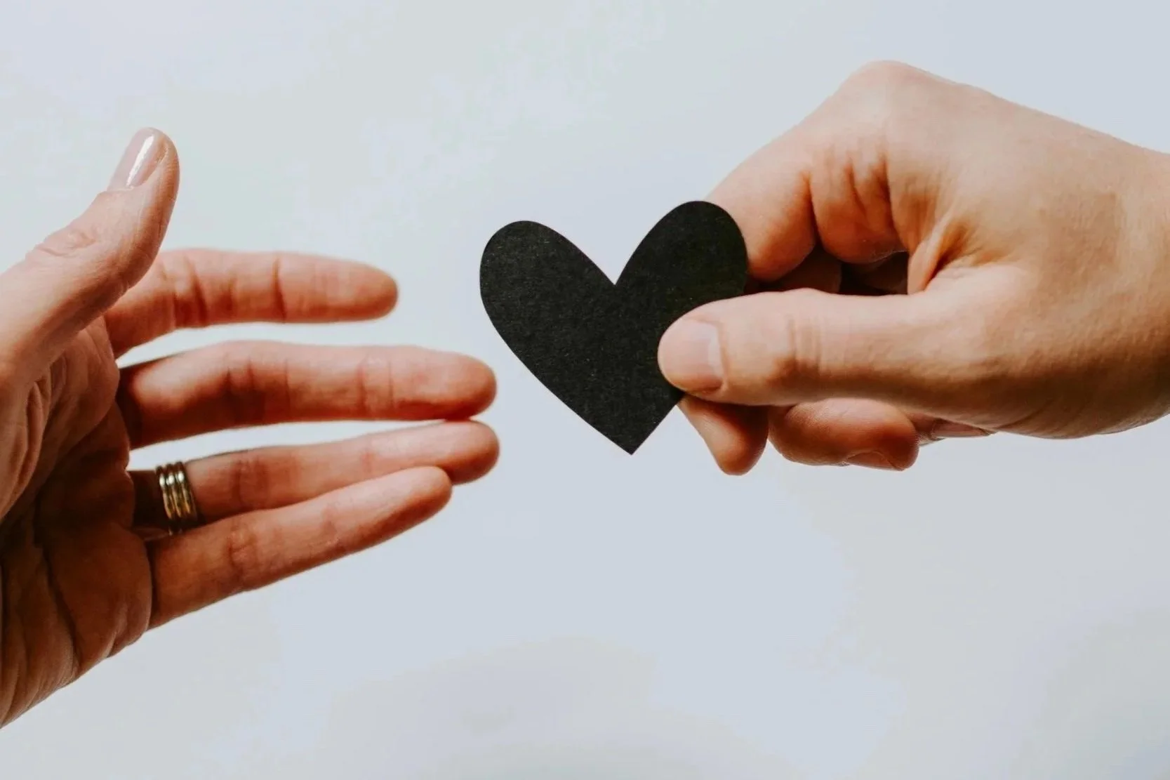 Deux mains, une tenant un cœur noir en papier et l'autre faisant un geste vers lui, sur un fond blanc.