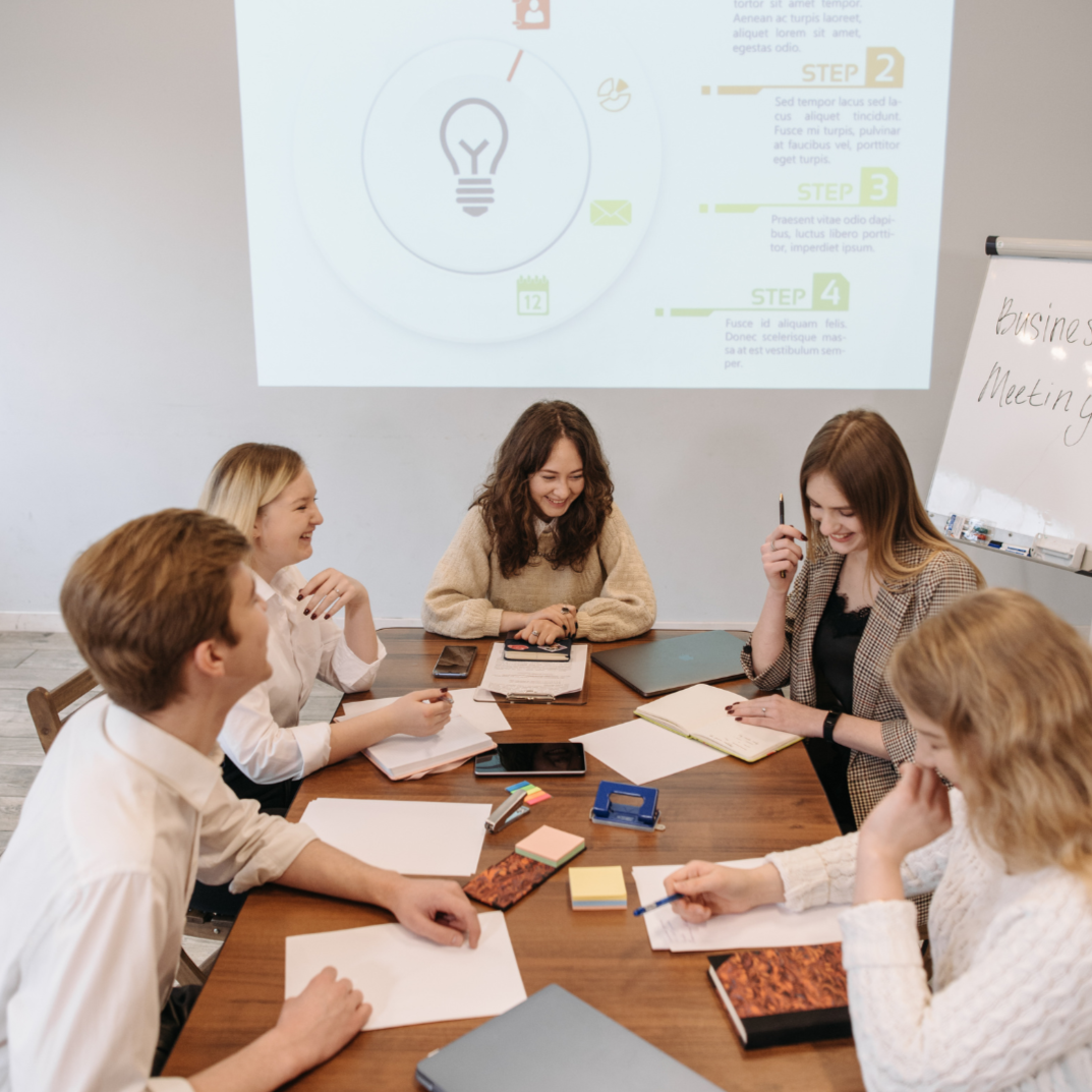 Six people are sitting around a conference table in a meeting room, smiling and discussing. There are notebooks, papers, and stationery on the table. A whiteboard with handwriting and a projected presentation are visible on the wall.