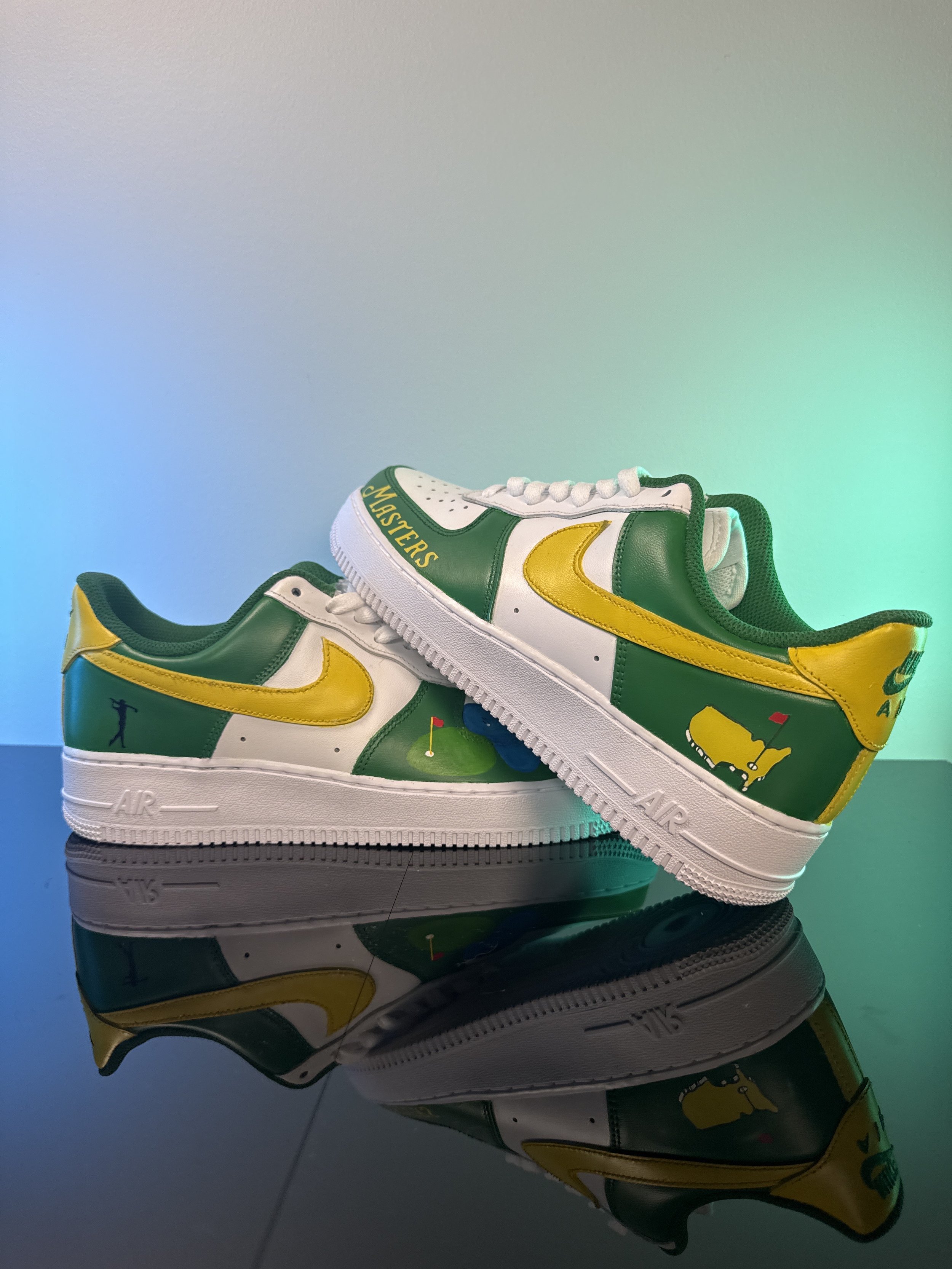 Pair of customized Nike Air Force 1 sneakers with golf-related artwork, green, yellow, and white coloring, displayed on a reflective surface with a colorful gradient background.
