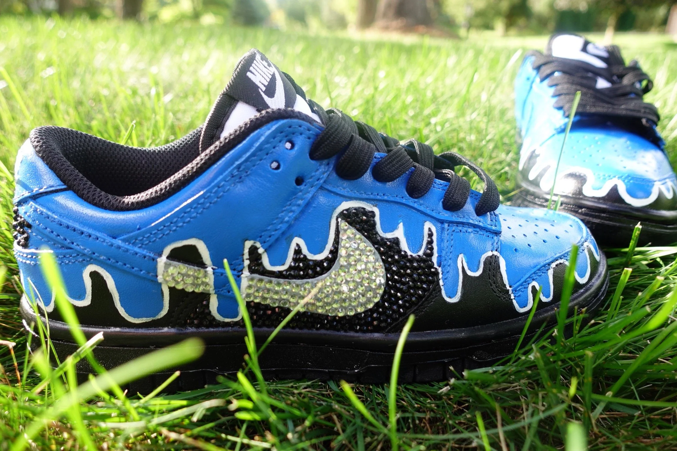 Blue and black Nike sneakers with rhinestone logo and decorative design, placed on grass outdoors.