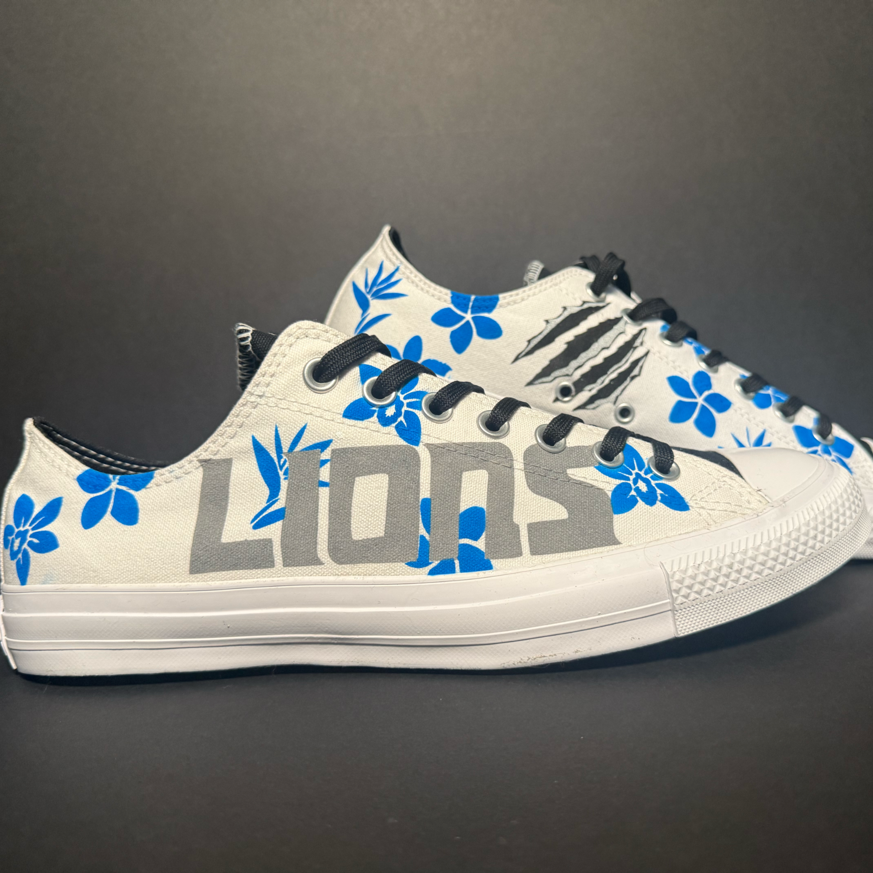 White low-top sneaker with a black heel tab and black laces, decorated with blue floral patterns and the word "LOVE" in large gray letters on the side, set against a dark background.
