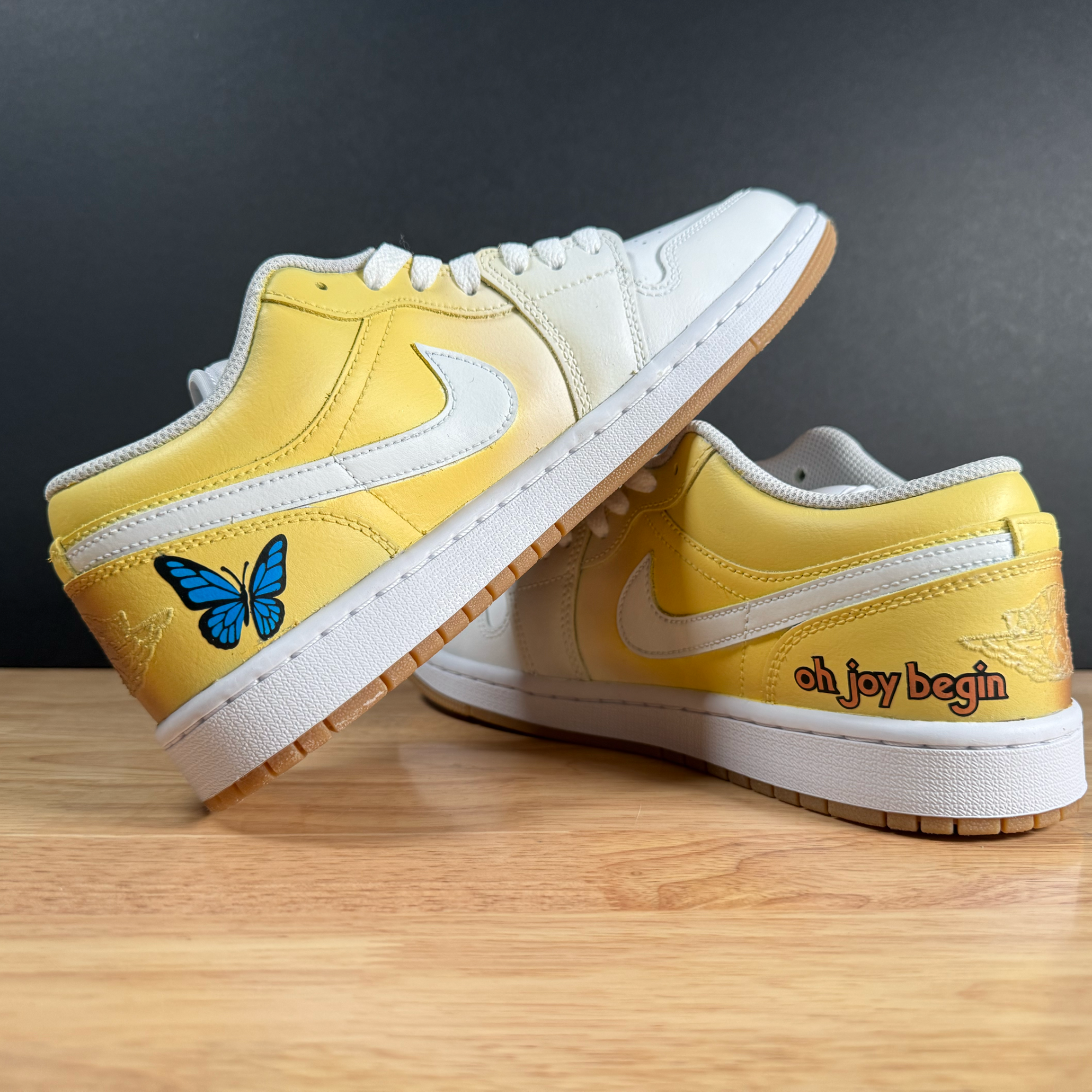 Close-up of a pair of yellow and white sneakers with a blue butterfly design on the side, placed on a wooden surface against a gray background.