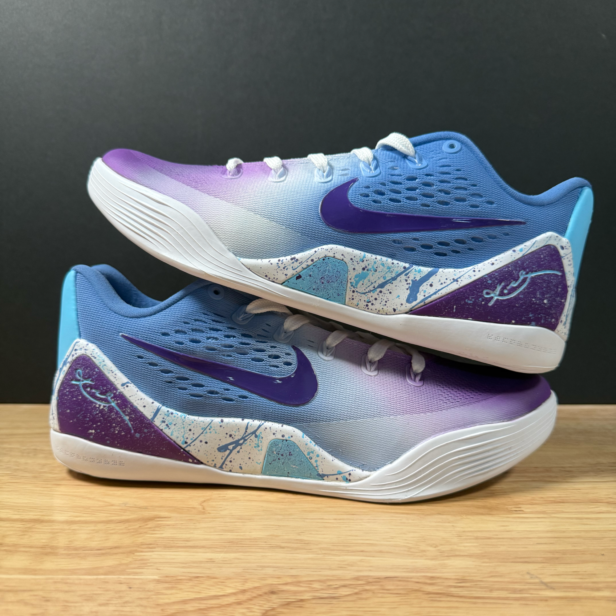 Close-up of a colorful running shoe with a purple and white gradient, purple Nike swoosh, and paint splatter design on the sole, placed on a wooden surface against a black background.