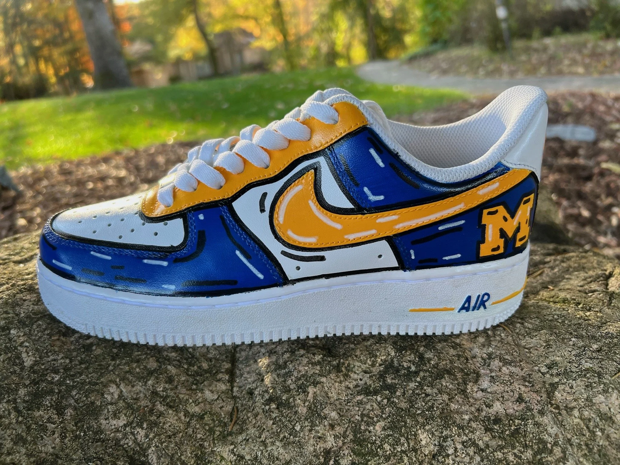 Custom painted white sneaker with blue, yellow, and black accents, featuring a Michigan Wolverines logo and the word 'AIR' on the sole, placed on a rock outdoors.