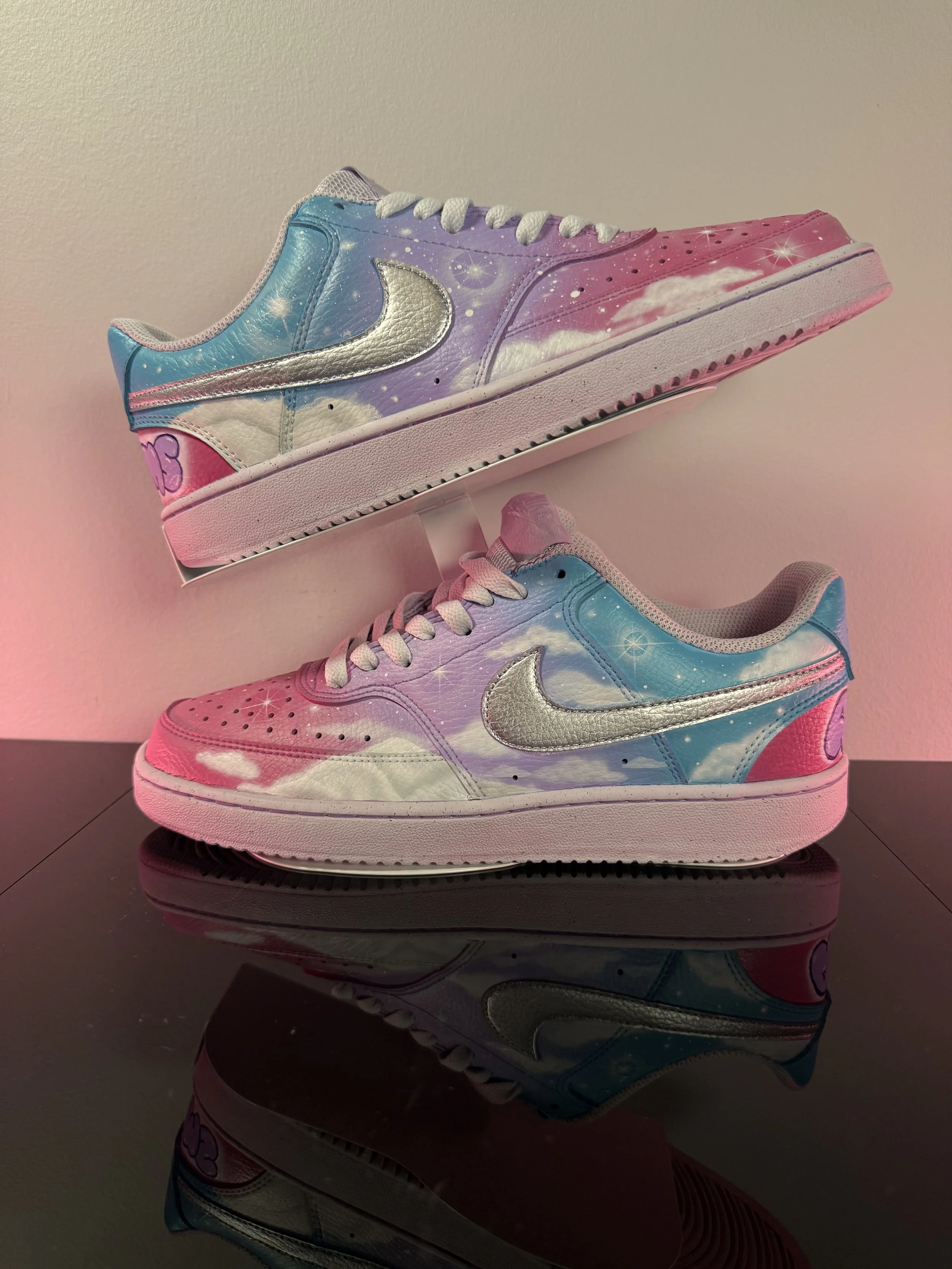 Pair of Nike sneakers with pastel galaxy design featuring clouds, stars, and the Nike logo, displayed on a reflective black surface.
