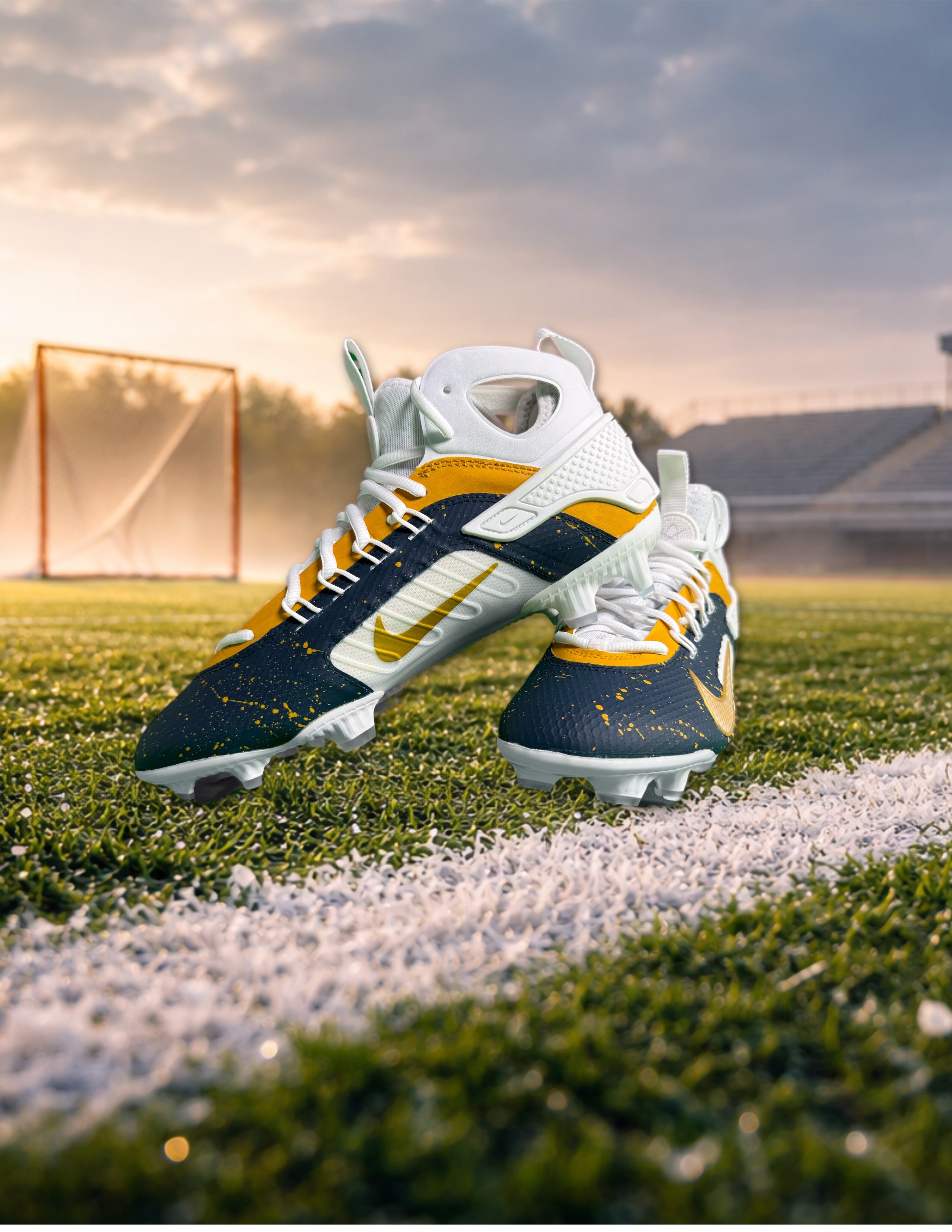 A pair of Nike football cleats in navy blue, white, and yellow on a football field at sunset.