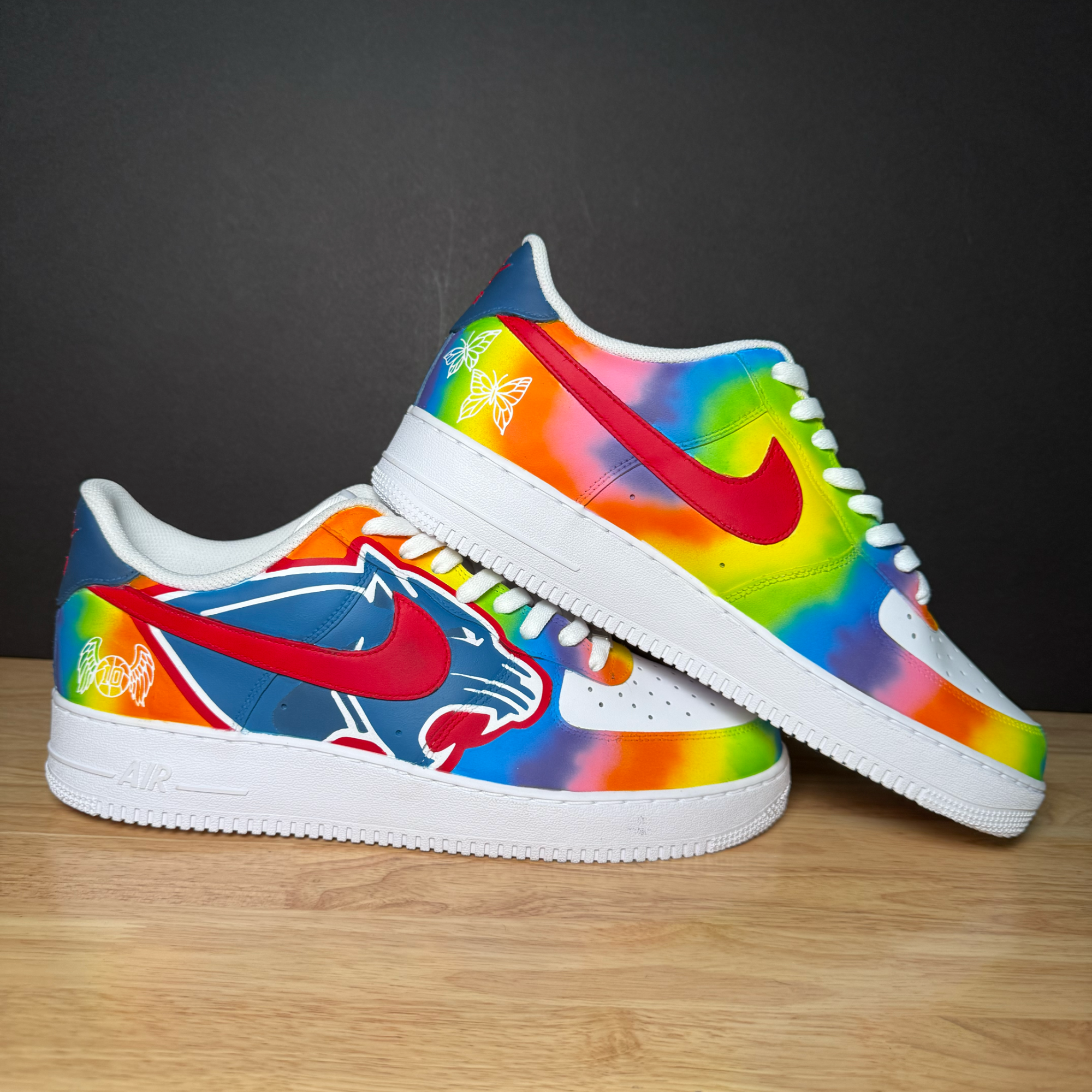 Colorful sneakers with rainbow gradient, red Nike swoosh, white soles, and butterfly and angel wing designs, placed on a wooden surface against a dark background.