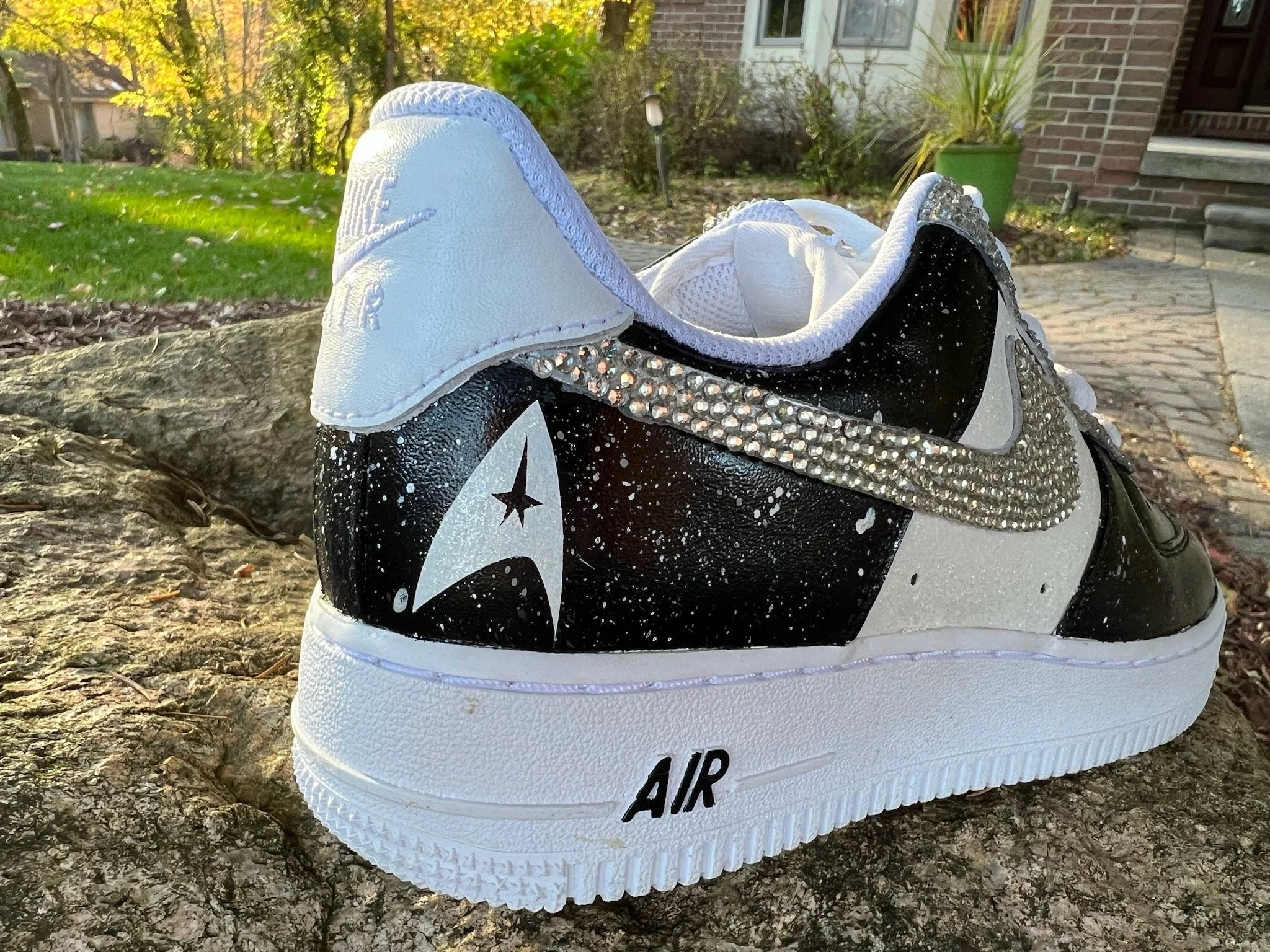 A sneaker with black, white, and silver colors, featuring rhinestones on the Nike swoosh, a star emblem, and the word 'AIR' on the sole, placed on a rock outdoors.