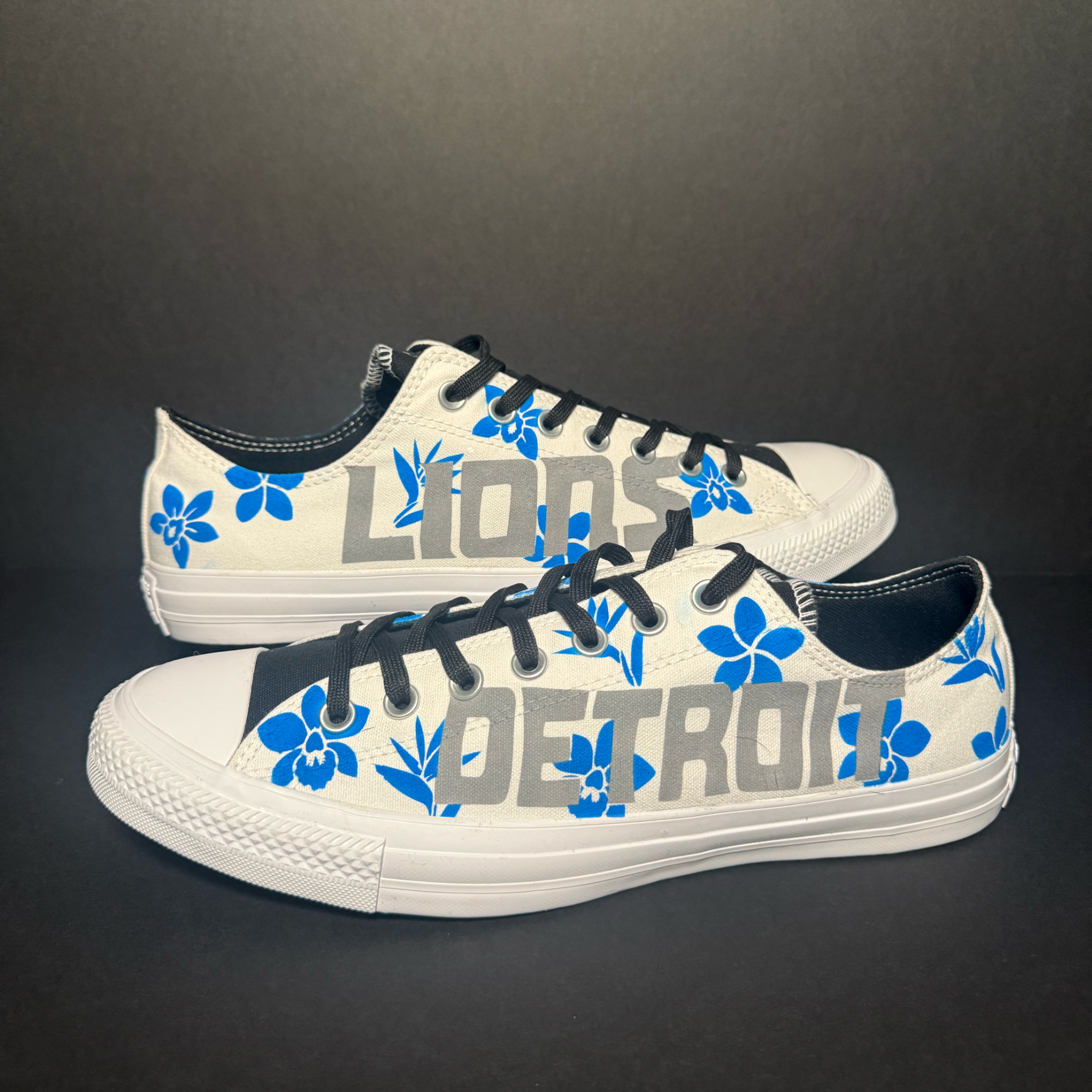 Close-up of a sneaker with white canvas, black laces, blue floral patterns, and gray lettering on a black background.