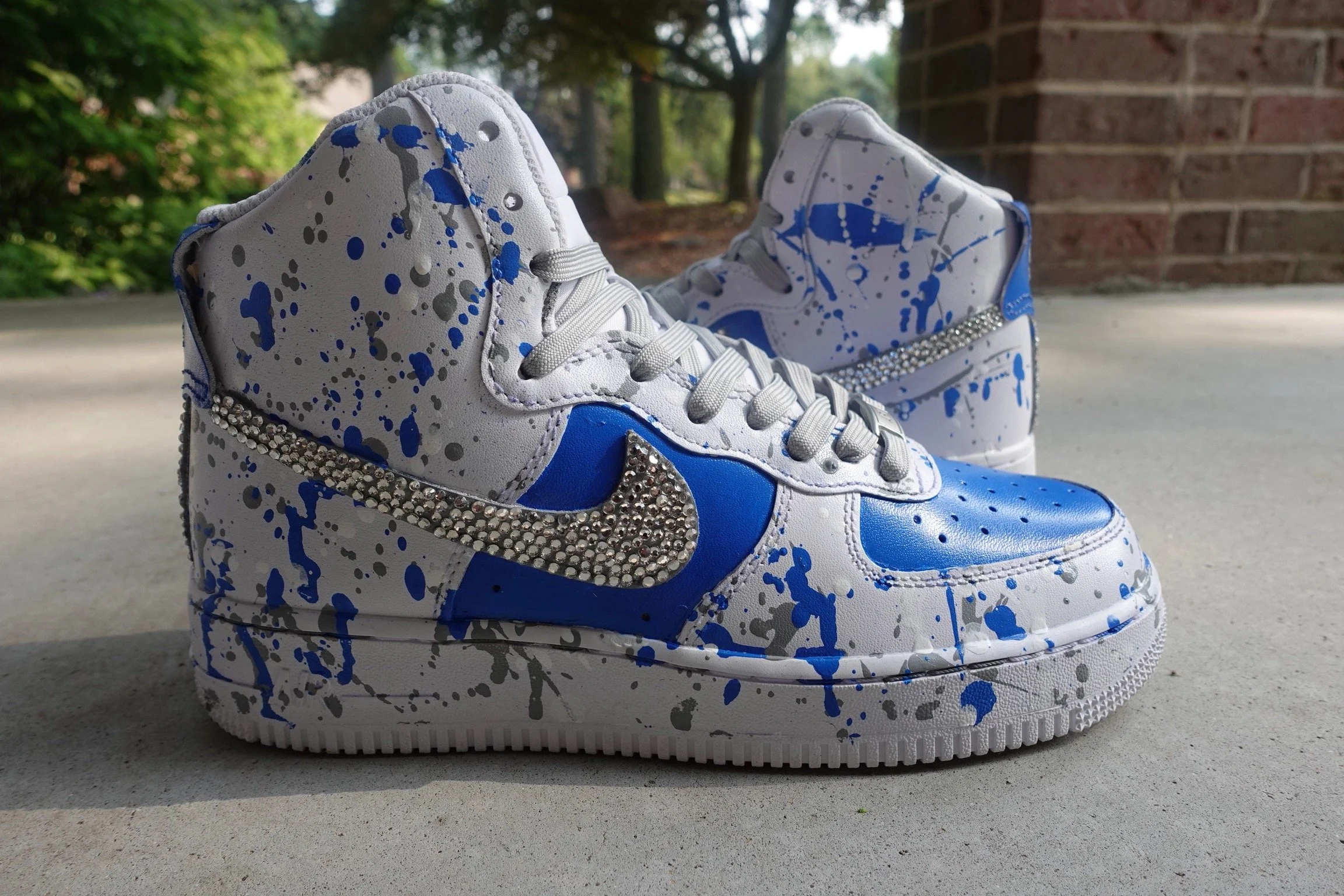 White sneakers with blue and gray paint splatter design, adorned with rhinestones on the Nike swoosh, outdoors on concrete with trees and brick wall in the background.