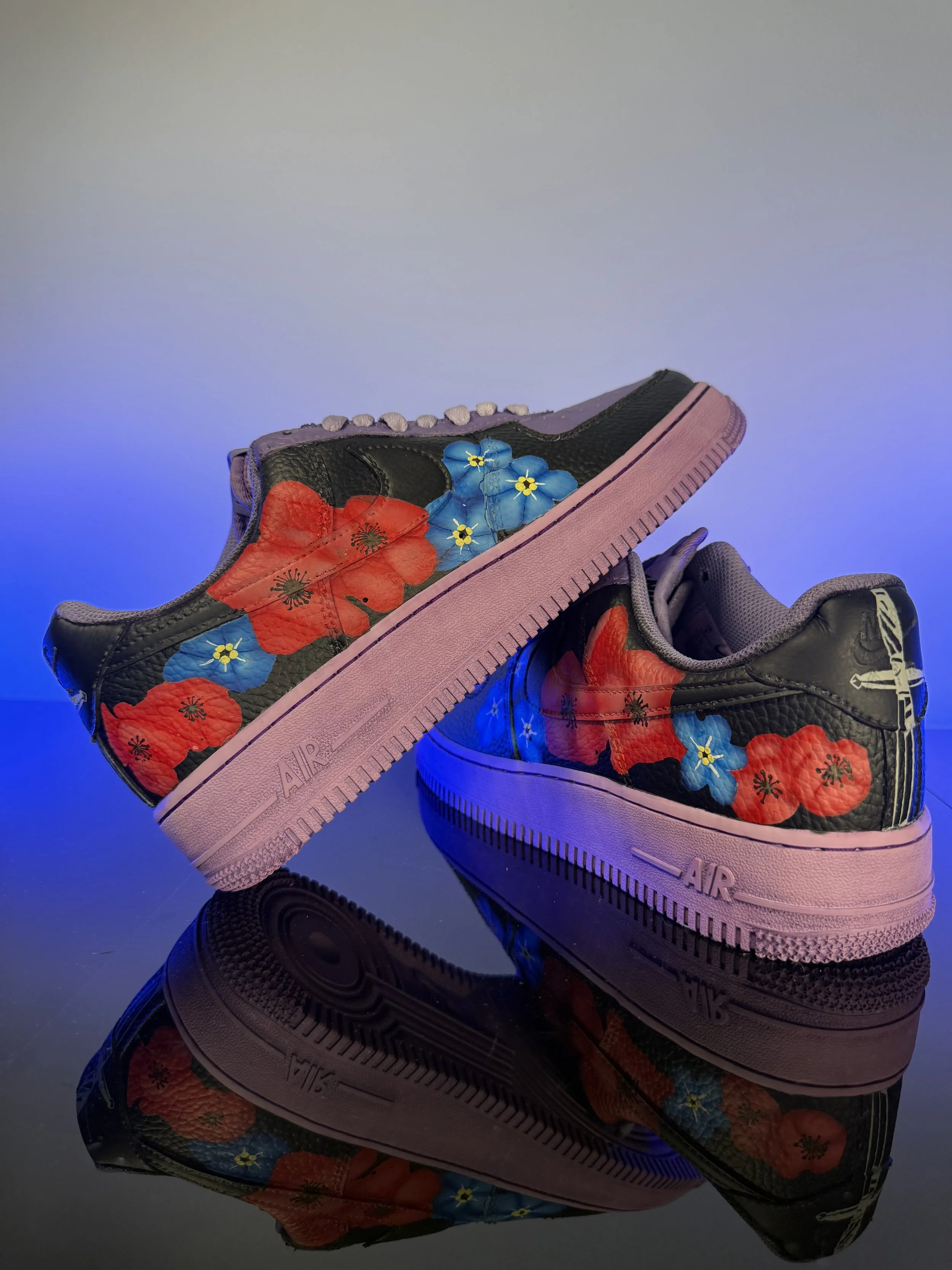 Pair of black sneakers with pink soles decorated with colorful floral designs, reflected on a black surface with a gradient background.