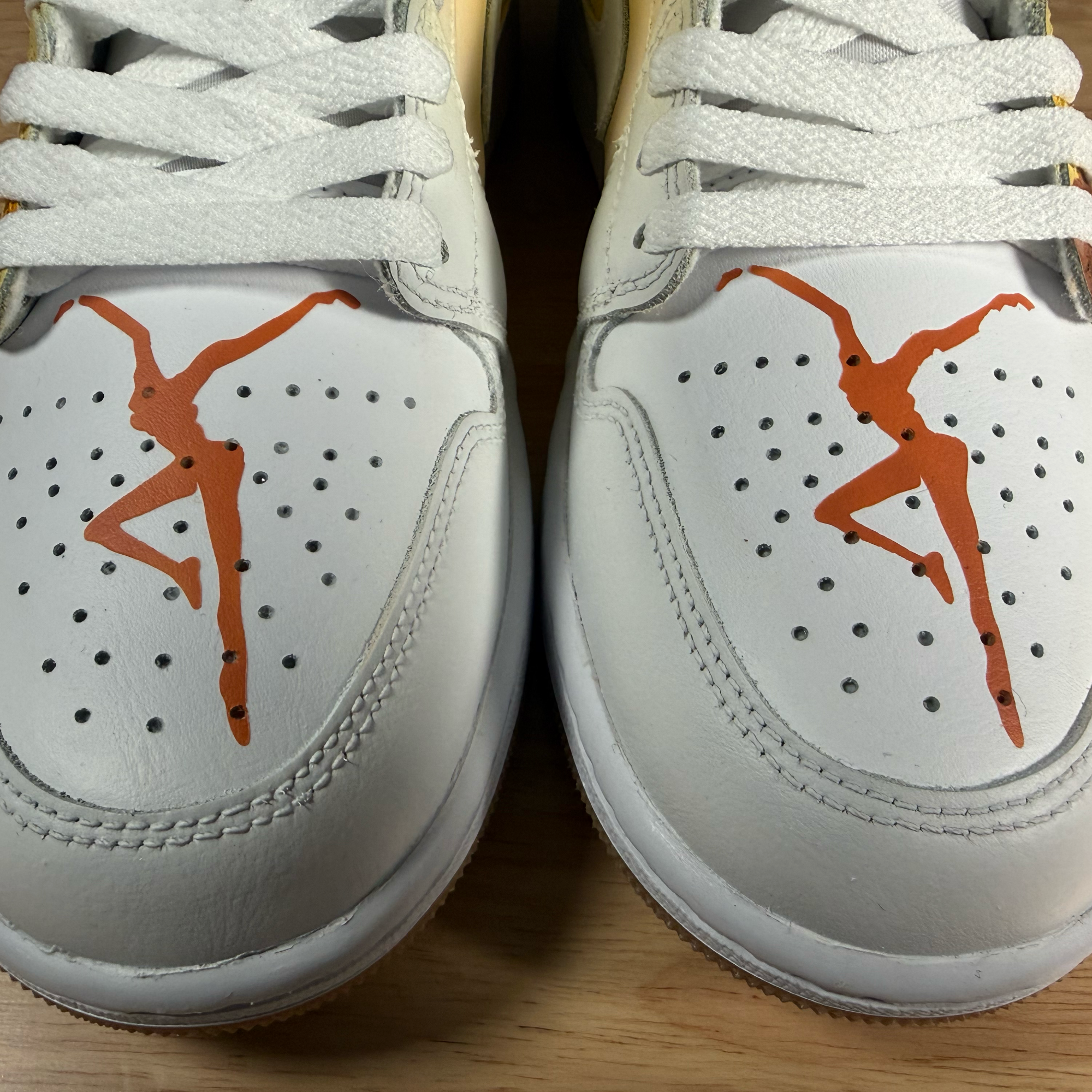 Close-up of white athletic shoes with orange Jumpman logos, showing the top and laces, placed on a wooden surface.