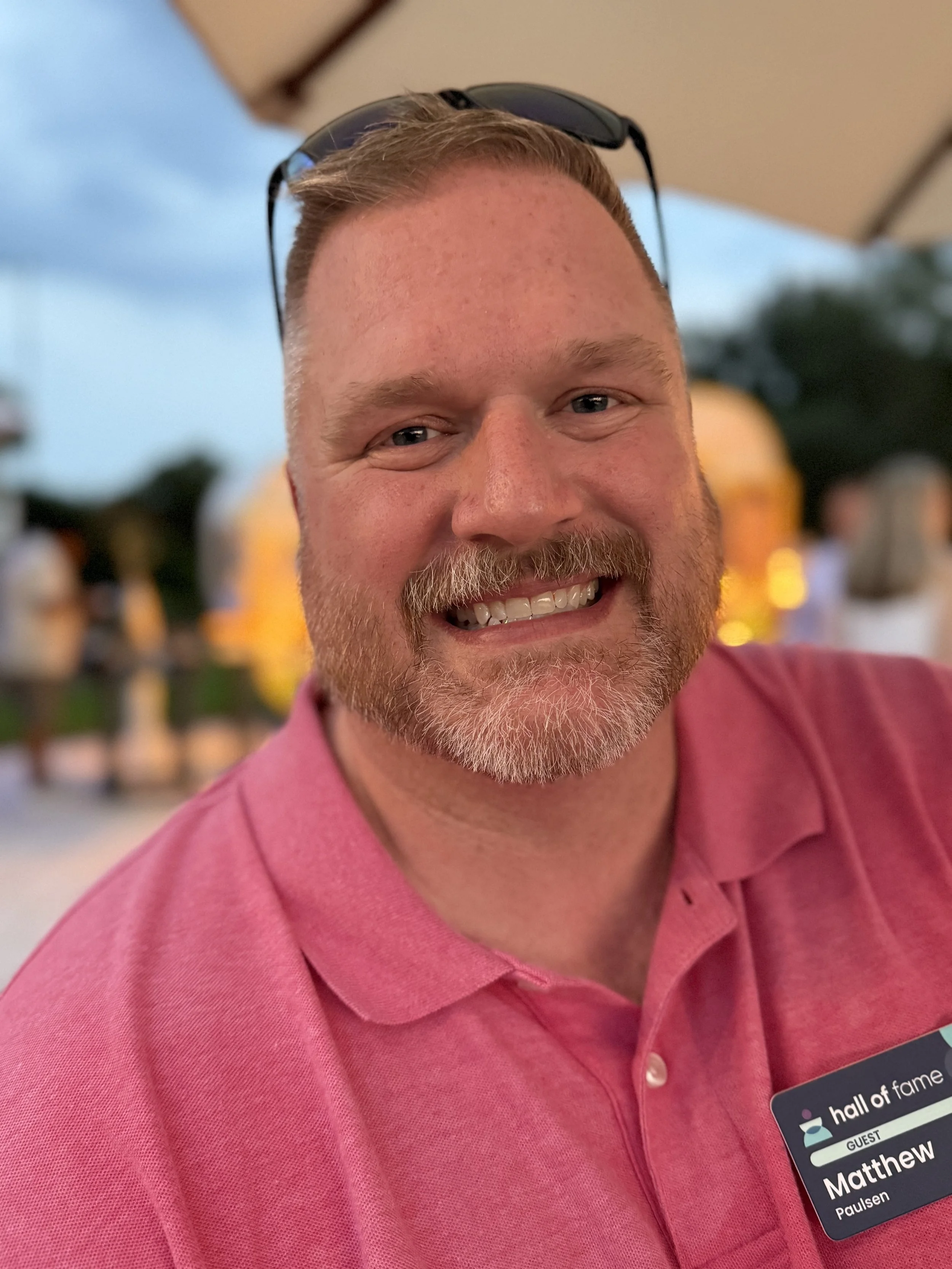 Close-up of a smiling man with a beard and mustache, wearing a pink polo shirt, with sunglasses resting on his head.