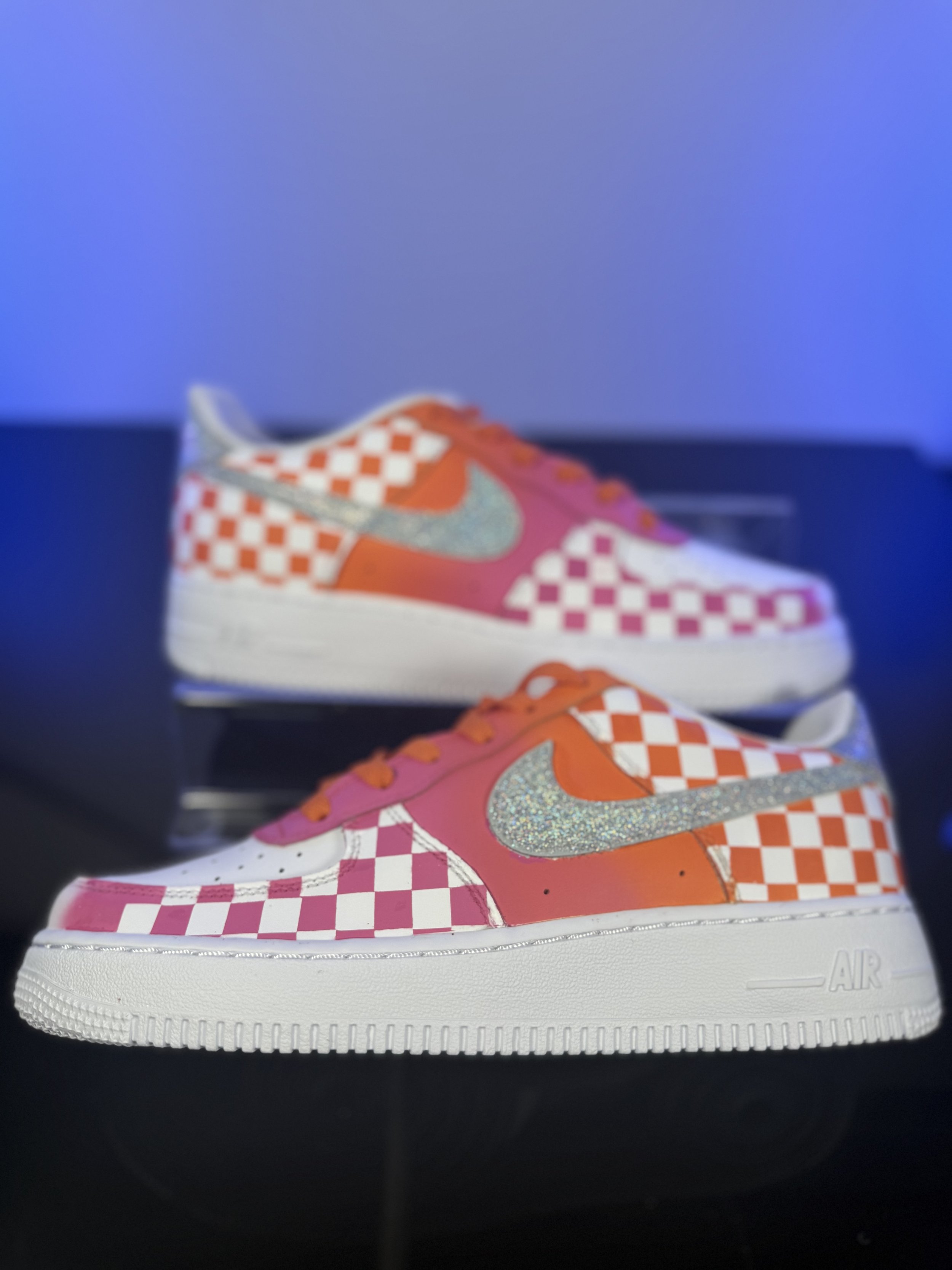 Colorful sneakers with orange, pink, white, and silver checkerboard and glitter details displayed on a black surface.
