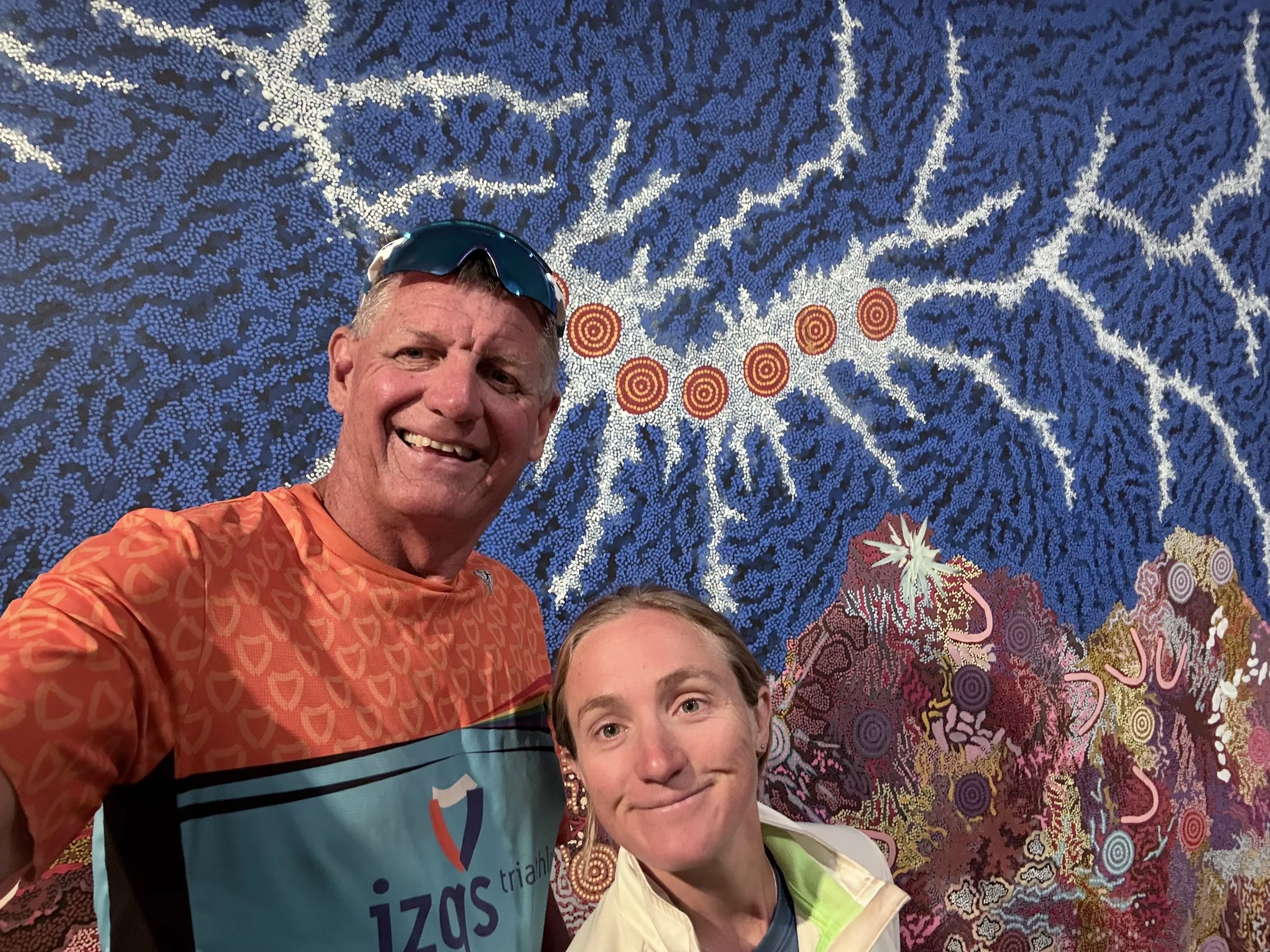 A smiling man and woman taking a selfie in front of an Aboriginal dot painting featuring white, blue, purple, and red patterns resembling a tree and roots.