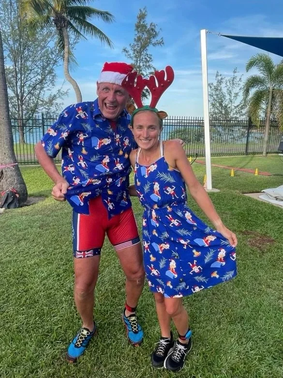 Two people outdoors in Christmas-themed attire, one wearing a Santa hat, reindeer antlers, and athletic shorts, the other wearing a dress with holiday print and reindeer antlers headband, smiling and posing for a photo.