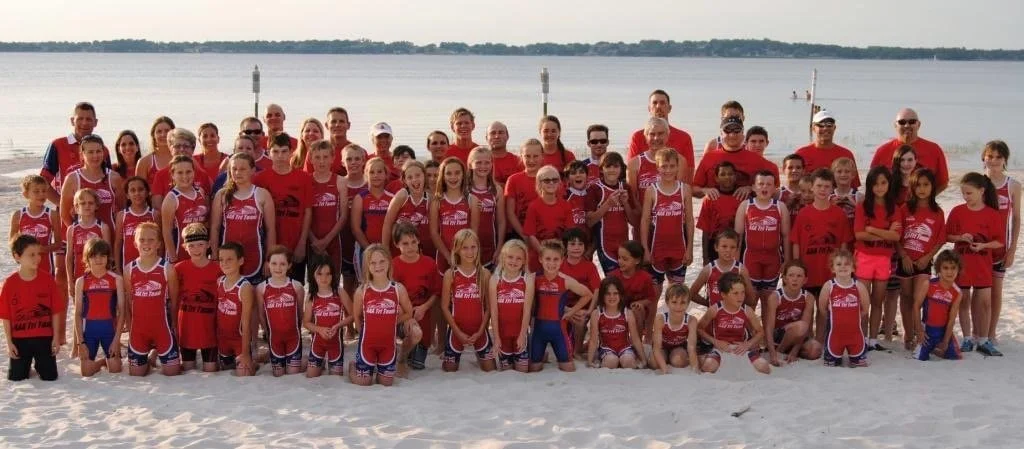 Group of children and adults in red athletic uniforms on a sandy beach by a body of water, possibly a team photo.