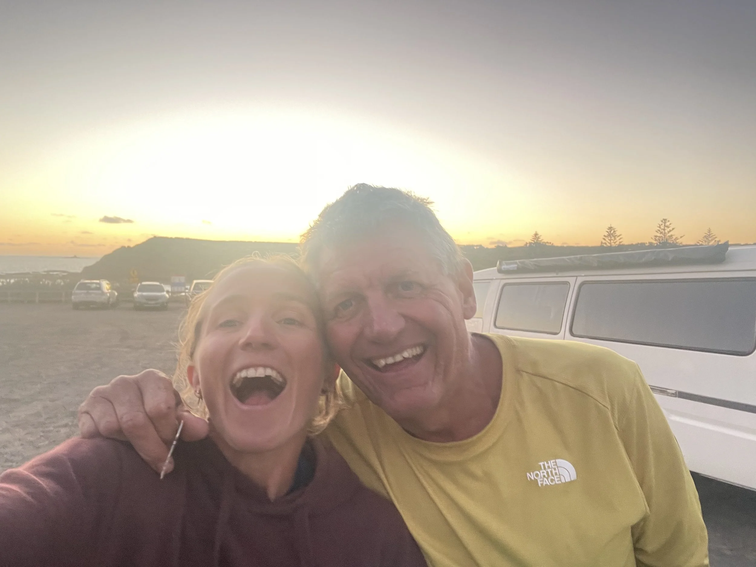 A happy couple taking a selfie at sunset on a beach with a vehicle and parked cars in the background.